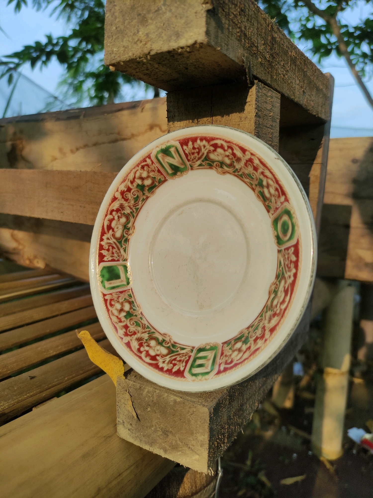 An overhead close-up shot shows a white, round, ceramic saucer with a decorative rim in red and green patterns and the letters "N", "L", "E", and "O" in green squares arranged around it. The saucer rests on a wooden structure with visible grain and texture, which is part of a larger wooden frame, possibly a bench or shelf, constructed from rough, weathered planks. The background is out of focus, showing dappled sunlight on more wooden surfaces, green foliage from trees, and a pale blue sky. The scene appears to be outdoors in a natural or rustic setting, possibly in Tiudan, Indonesia, given the context. The lighting suggests it is daytime, with shadows cast by the wooden structure. No people are present in the image.