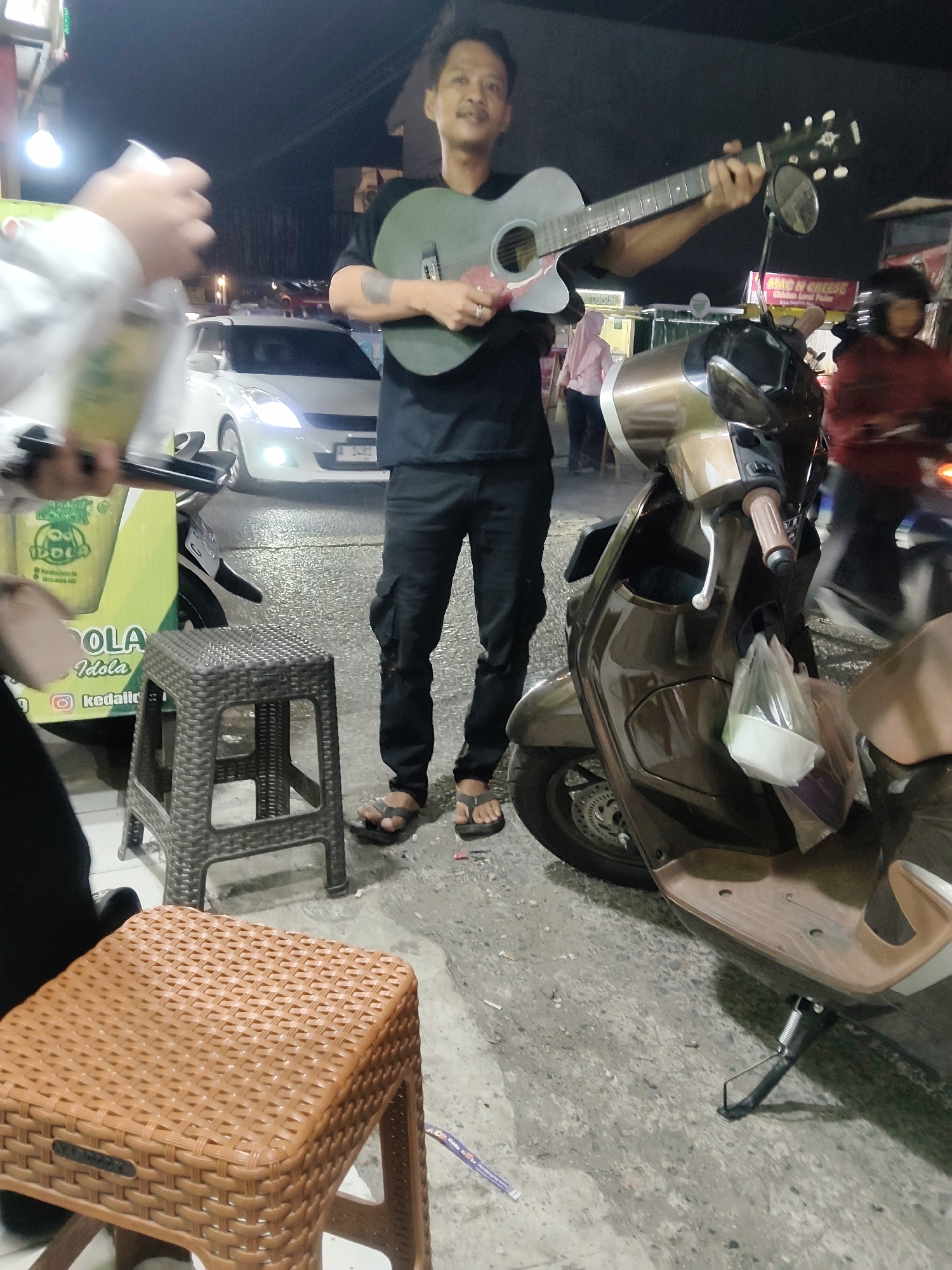 In Mekarmukti, Indonesia, a man stands on a paved street at night, playing an acoustic guitar. He is wearing a black t-shirt and black cargo pants, with sandals on his feet. The guitar is light green and he is holding it with both hands. To his right, a brown scooter is parked, with a plastic bag hanging from its handlebars. In the background, a white car is visible, along with other people and a food stall. Two plastic stools, one dark gray and one brown, are placed in front of the man. The overall scene suggests a street performance or casual gathering in a public space.