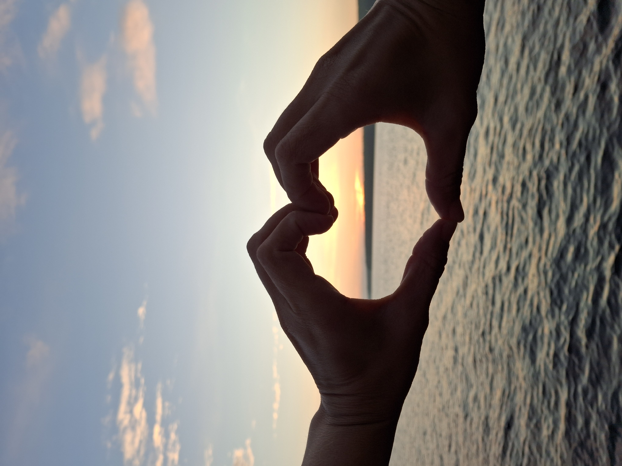In Chaikovsky, Russia, two hands form a heart shape against a sunset sky over a body of water. The sky is a gradient of blue and orange, with scattered clouds. The water reflects the colors of the sky and appears slightly rippled. The hands are silhouetted against the light, obscuring any distinguishing features. The scene evokes a sense of romance or affection.