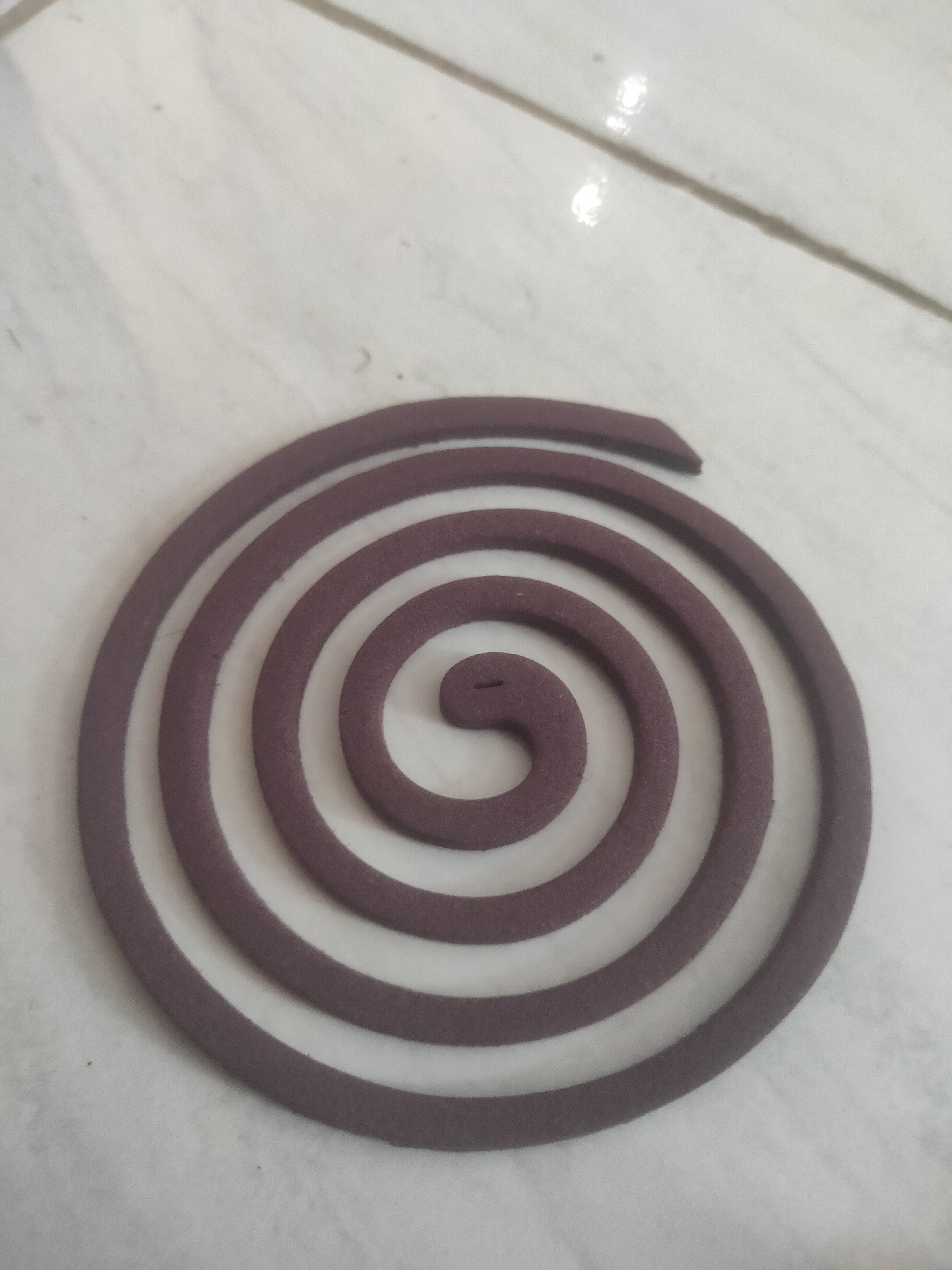 An overhead shot shows a dark purple mosquito coil resting on a pale, tiled surface. The coil is wound in a spiral pattern, with several complete rotations visible. The tiled surface appears to be a floor, with faint lines indicating grout between the tiles. The lighting suggests an indoor environment, possibly a home or an outdoor covered area in Antirogo, Indonesia. There are no people or other objects in the scene. The focus is entirely on the mosquito coil.