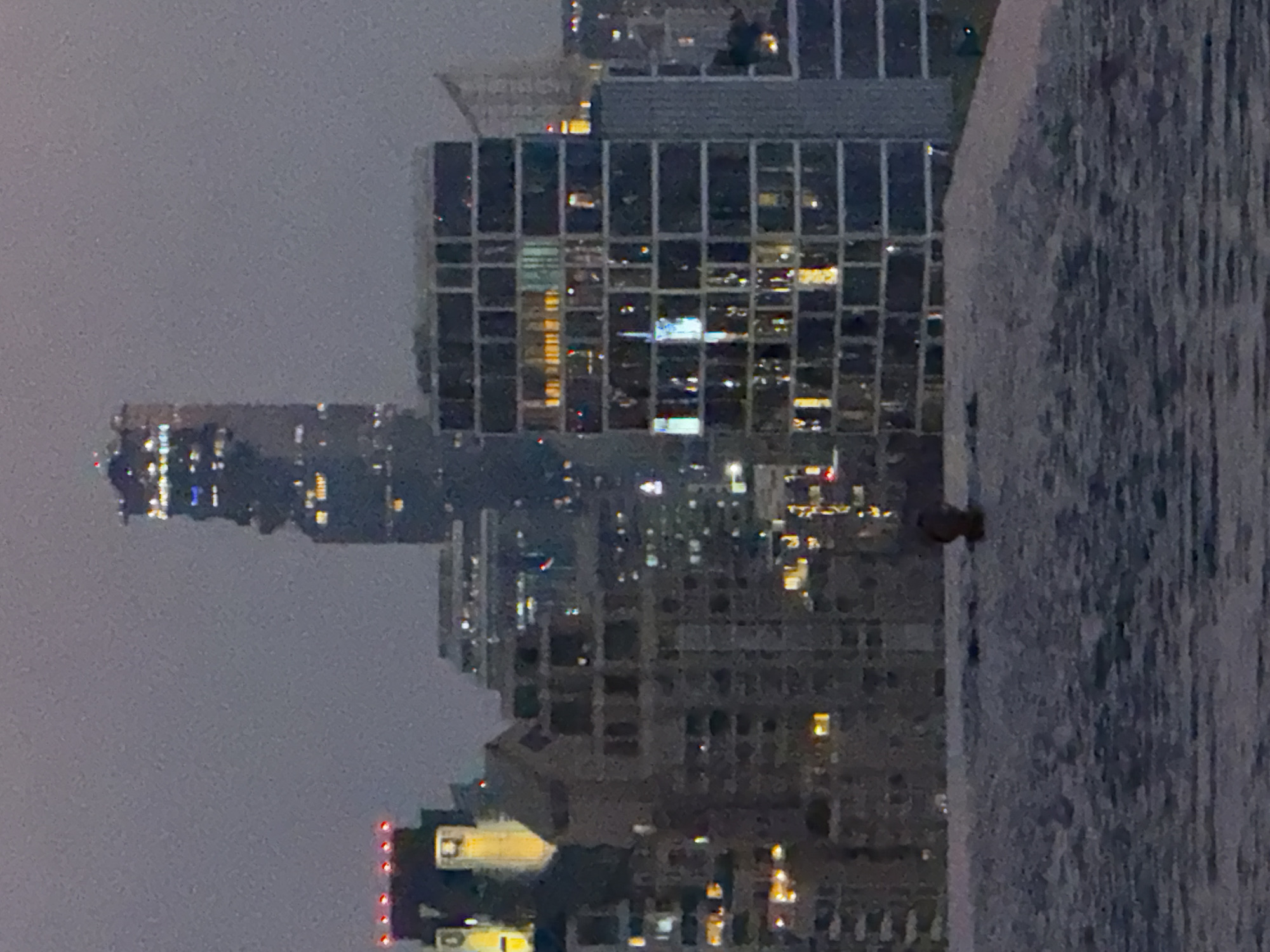 The image shows a nighttime view of the Bangkok skyline from a high vantage point. Several illuminated skyscrapers are visible, with one prominent building featuring a grid-like pattern of windows. The sky is overcast. A body of water, possibly a river or swimming pool, is in the foreground. A single person is in the pool.