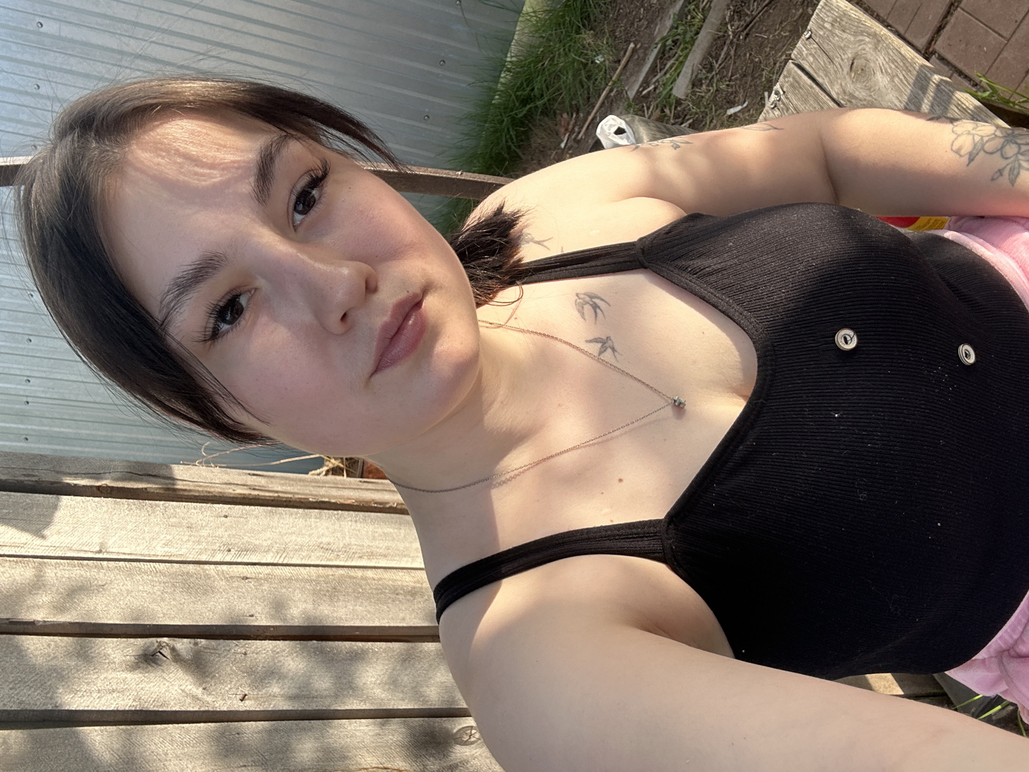 A fair-skinned woman with dark hair is lying on a wooden surface outdoors, likely a bench or deck, in what appears to be sunlight. She is wearing a black tank top with two metal buttons on the front and a delicate silver necklace with a pendant. Her hair is pulled back, and she has well-defined eyebrows and eyelashes. She is looking directly at the camera. The woman has tattoos on her chest and arms; the chest tattoo appears to be birds in flight.
The background includes a galvanized metal fence, green foliage, and more wooden planks. The location is not explicitly specified in the city of Chaikovsky, Russia, though the user has requested the details.