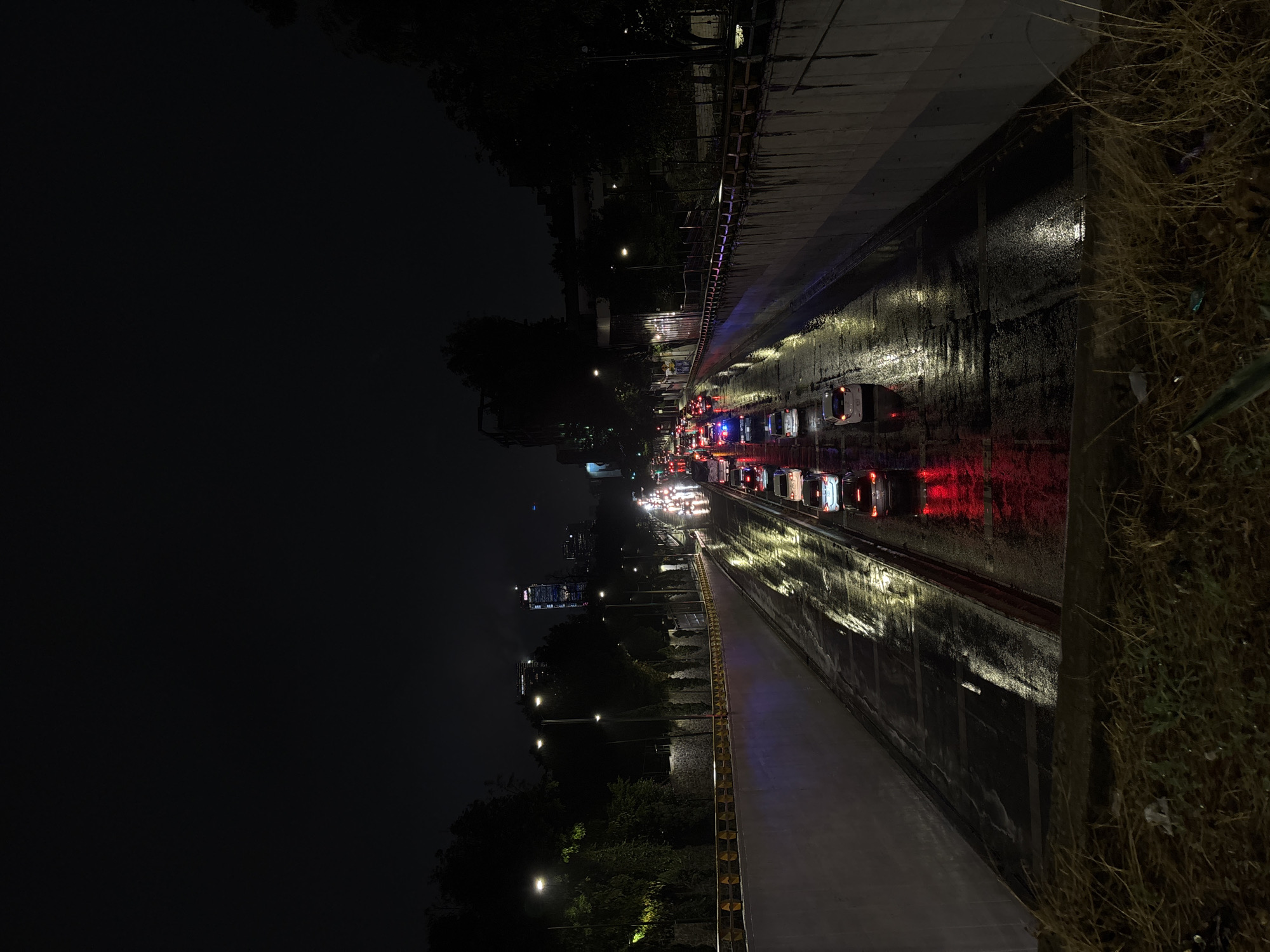 The image depicts a nighttime view of a wet, multi-lane road in Mexico City, Mexico. Traffic is visible, with the red taillights of numerous vehicles indicating a congested road. Streetlights and vehicle headlights create bright reflections on the wet pavement, while buildings can be seen in the background. A concrete barrier runs along the side of the road, with grass and other vegetation growing adjacent to it. The left side of the image appears dark, suggesting a lack of artificial lighting in that area.