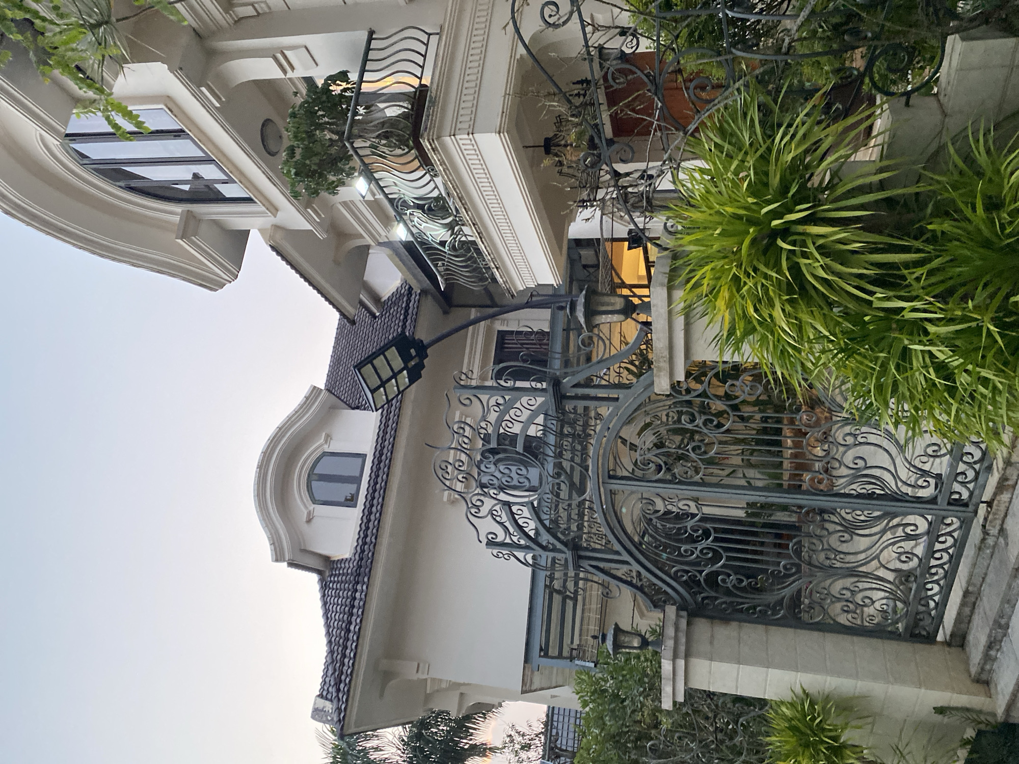 A low-angle view of a multi-story building with a beige exterior, situated in Da Lat, Vietnam. The building features balconies with ornate wrought-iron railings and decorative moldings. A gray wrought-iron gate with elaborate scrollwork stands at the entrance. A streetlight with a modern rectangular lamp head extends over the gate. Lush green plants and trees flank the gate and obscure parts of the building. The sky is a pale gradient, suggesting either dawn or dusk.