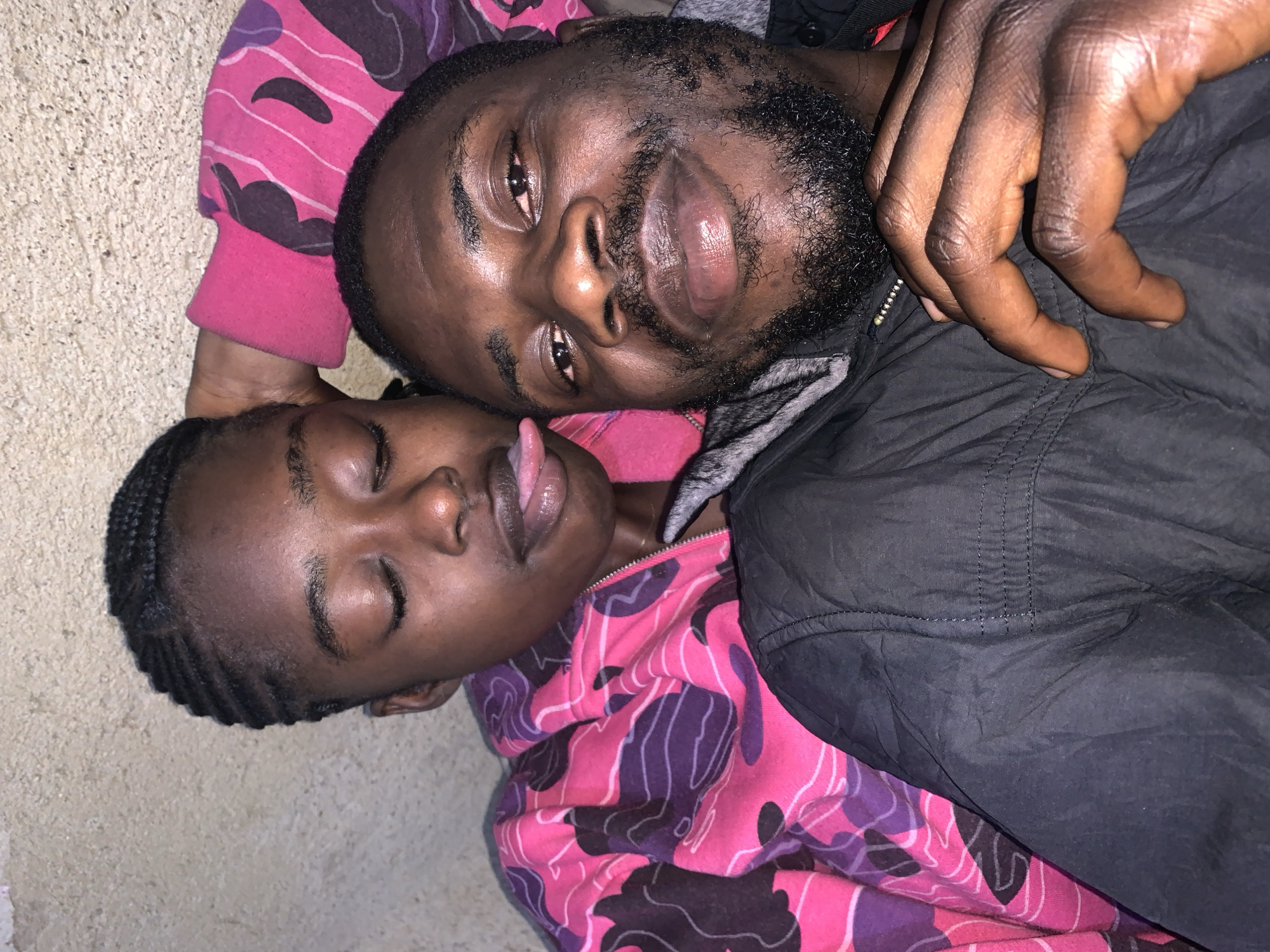 The image depicts two individuals, an adult male and a young female, in a close embrace. The background is a textured, light-colored wall, possibly an exterior surface, consistent with locations in Rafin Par, Nigeria.
The young female, positioned in the foreground and to the left, has her eyes closed and her tongue sticking out. She is wearing a vibrant pink and purple camouflage-patterned hooded jacket. Her hair is styled in braids.
The adult male is to the right, his face angled towards the camera. He has dark skin, short, dark hair, and a beard. He appears to be wearing a black jacket. His right arm is wrapped around the young female, and his hand rests on her shoulder. His left hand is visible in the lower right quadrant of the image, fingers curled. The lighting suggests an indoor or evening setting, with light reflecting off their skin and lips. The overall impression is one of intimacy and comfort between the two subjects.
