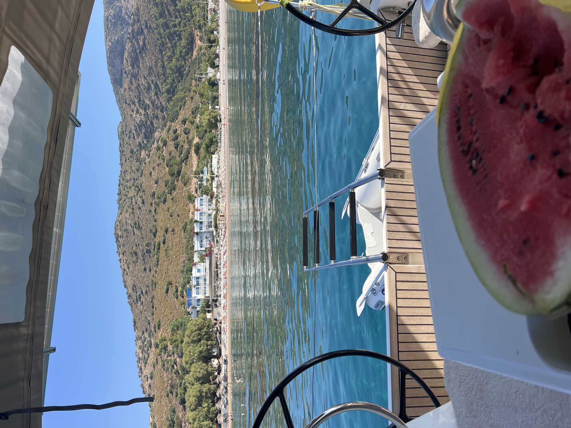 The image captures a view from the deck of a boat, looking out towards a coastline. In the foreground, a portion of a watermelon is visible, slightly out of focus, placed on a white surface. To the left, a dark-colored steering wheel of the boat is prominent. The boat deck is made of polished wooden planks. In the middle ground, a white inflatable dinghy with a small outboard motor is attached to the stern of the boat, with a stainless steel ladder extending from it. The water is a clear turquoise, with subtle ripples on its surface. Beyond the water, a sandy beach lined with numerous beach umbrellas and lounge chairs stretches along the shore. Several people are visible on the beach and in the shallow water. Behind the beach, a low, tree-covered hill rises, dotted with vegetation. Further back, a multi-story white hotel with blue trim is situated on the hillside overlooking the beach. The sky is a clear, bright blue, indicating a sunny day. The scene appears to be in a coastal resort area.
