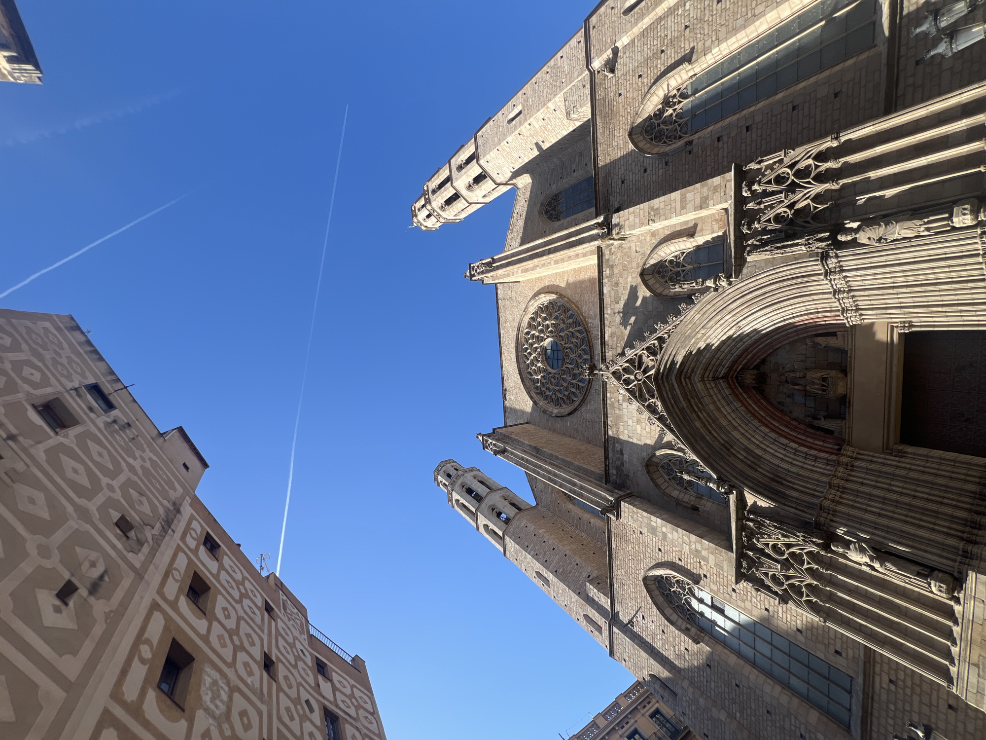 The image displays a low-angle view looking directly upwards at the Basilica de Santa Maria del Mar in Barcelona, Spain. The central subject is the grand, light-brown stone façade of the Gothic cathedral, illuminated by direct sunlight which casts distinct shadows and highlights the architectural details.
The cathedral's main entrance features a deeply recessed, pointed archway adorned with intricate carvings and statuary within niches. Above the entrance, a large, ornate rose window with delicate tracery is prominently centered. The façade is flanked by two tall, octagonal bell towers, each with multiple levels of arched openings. The stone surface of the building exhibits weathered textures and variations in tone. Along the sides, supporting structures and tall, slender stained-glass windows with pointed arches are visible.
The upper portion of the frame is dominated by a clear, vibrant blue sky. Two white airplane contrails traverse the sky: one faint and straight, extending diagonally from the lower-left sky to the upper-right, and another, brighter contrail also running diagonally across the upper-right quadrant.
In the upper-left corner, a portion of an adjacent building's façade is visible, distinguished by a geometric pattern of alternating light beige and off-white diamond and square motifs. Small, dark window openings are interspersed within this pattern. A less distinct portion of another building is visible in the upper-right corner. There are no people, vehicles, or discernible actions within the frame. The scene conveys a sense of architectural grandeur under a bright, open sky.