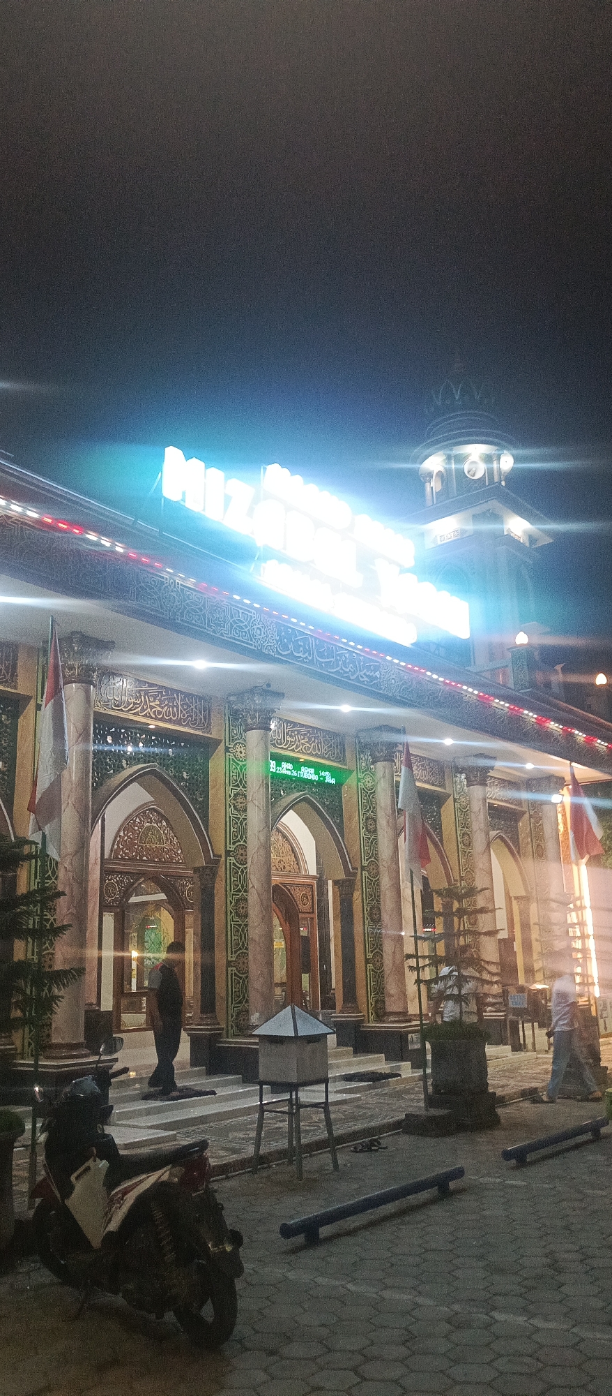The image depicts the entrance of a mosque in Arjasa, Indonesia, at night. A prominent neon sign, illuminated in bright white and blue, displays text that is partially obscured but appears to include the word "Masjid". The mosque's facade features ornate columns with marble detailing and Arabic calligraphy. Two Indonesian flags are mounted on poles near the entrance. In the foreground, a motorcycle is parked on a paved surface made of interlocking bricks. A few individuals are visible: one person stands near the mosque entrance, and another is seen walking further inside. A digital display showing a date and time is mounted on the wall. The overall environment suggests a public space within the city.