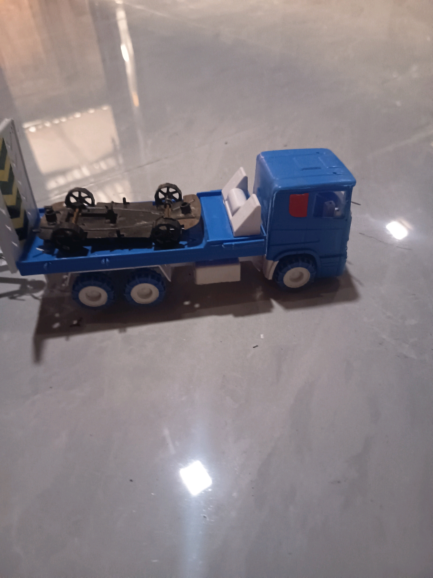 A blue toy truck is carrying a small, dark-colored toy car on its flatbed. The car is upside down. The truck has six white wheels with yellow rims. The truck's cab is blue with a red window. The rear of the truck is yellow and black striped. The toys are on a gray, reflective surface. The image appears to have been taken indoors. The specific location within Sidorejo, Indonesia, is not identifiable from the visual information provided.