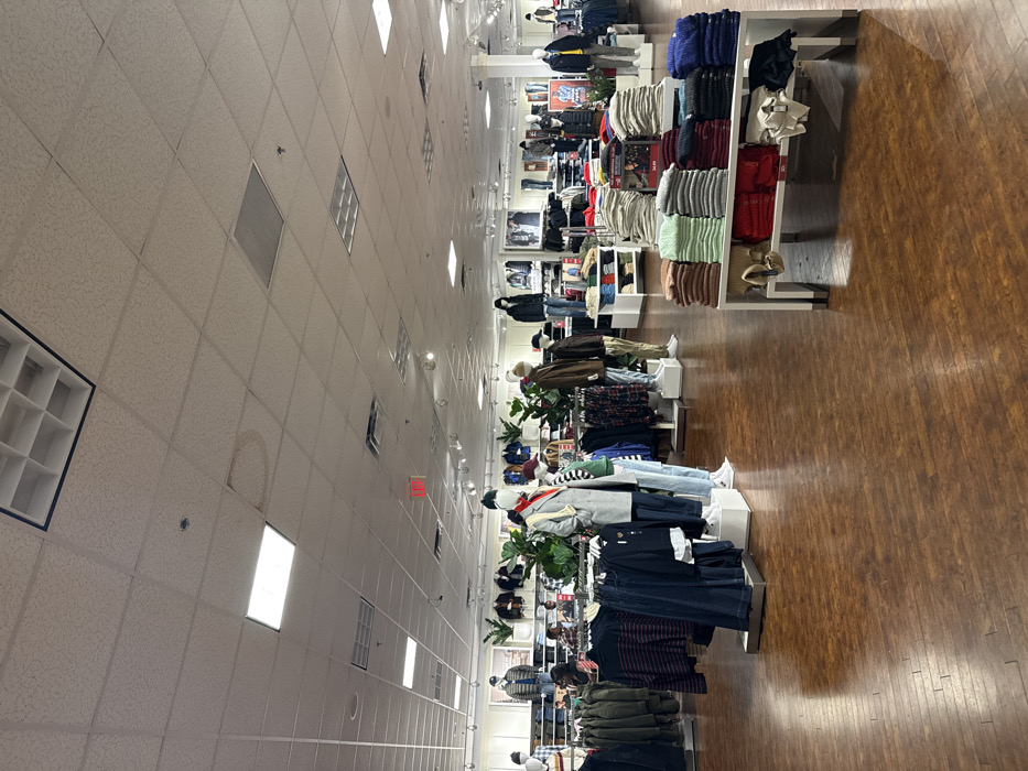 The image shows the interior of a clothing store. Mannequins dressed in various outfits stand in a line, positioned between rows of folded garments on shelves. The floor is made of wood planks. Overhead, a ceiling comprised of square tiles is visible, with various light fixtures and ventilation panels. The store appears well-lit and organized.