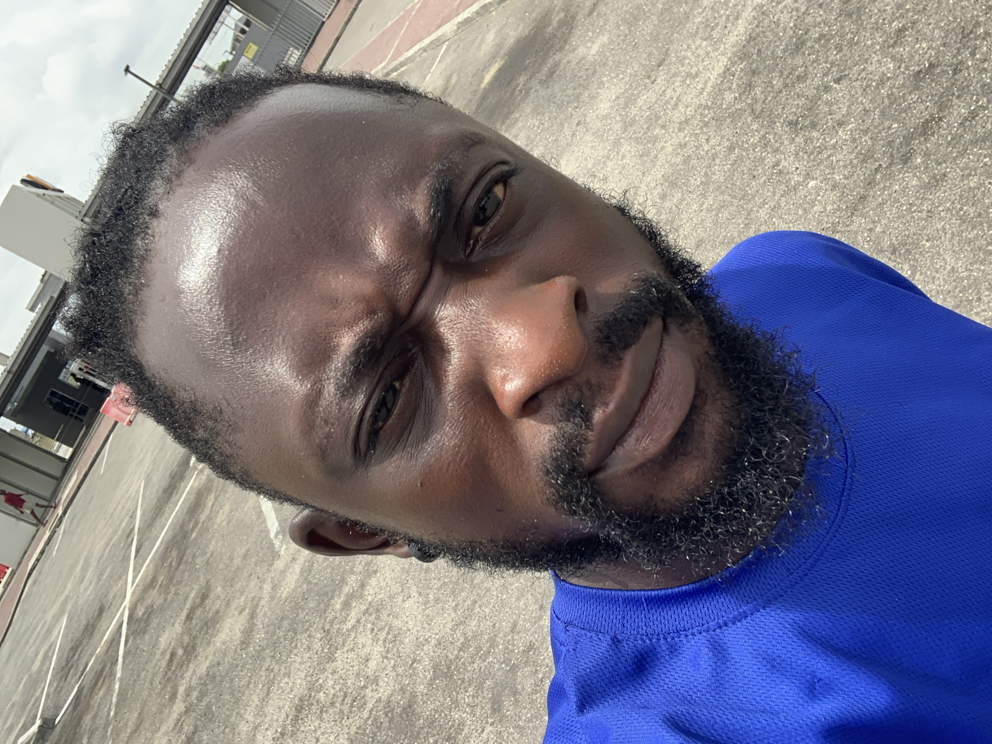 A medium, close-up shot shows a Black man wearing a blue shirt. His expression is intense, with his brow furrowed. He has a full beard and short hair. The background shows an asphalt surface with parking lines, and a building complex with metal structures. The location is possibly Sangotedo, Nigeria, given the context.