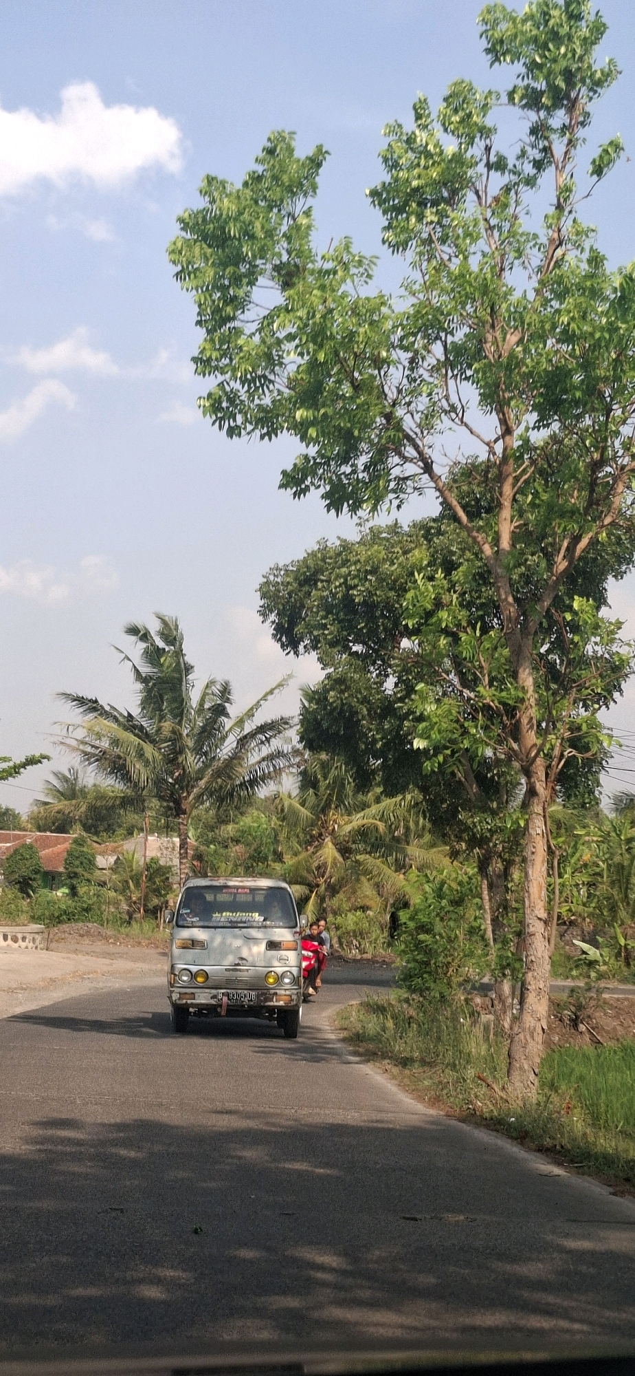 In the city of Cibinong Hilir, Indonesia, a light-colored van is driving on a paved road under a clear sky with scattered clouds. The van is silver and appears to be a commercial vehicle. Two people are riding on a motorcycle behind the van, with one person driving and the other as a passenger wearing a red shirt. The road is lined with lush green trees and vegetation, including palm trees, typical of a tropical environment. In the background, houses with red roofs are visible amongst the trees, suggesting a rural or semi-rural setting. The sun is casting shadows on the road, indicating it is daytime. The scene conveys a sense of everyday life in a peaceful Indonesian town.