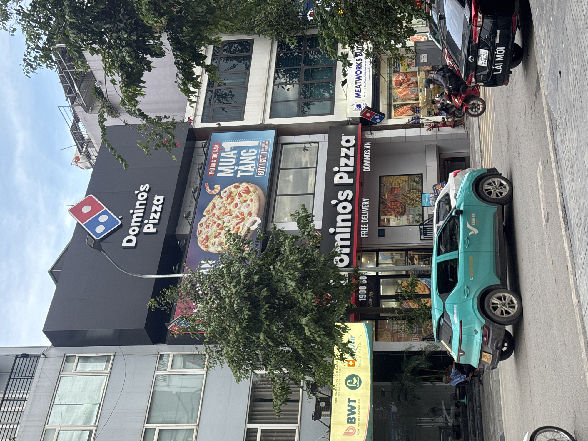 This is a medium shot of a street scene in Hà Nội, Vietnam, captured on a cloudy day. In the foreground, a teal-colored electric car, likely a VinFast VF e34, is parked on the right side of the road, facing left. To its right, a black sedan with a Vietnamese license plate is also parked. Behind these vehicles, several businesses are visible. A prominent Domino's Pizza facade is centered, with its logo and "Domino's Pizza" signage clearly displayed. Adjacent to it, a shop labeled "Meatworks Butcher" is also present. Further to the left, a yellow banner displays the BWT logo and "BEST WATER TECHNOLOGY." A tree with dense green foliage stands in front of the Domino's Pizza establishment, partially obscuring the view of the building. People are sparsely visible; one individual sits on a chair outside a shop to the left, and a person on a red motorcycle is seen in the middle ground. The street is paved, and a pedestrian crossing is marked with yellow lines. The architecture of the buildings appears to be modern, with multiple stories and large windows.