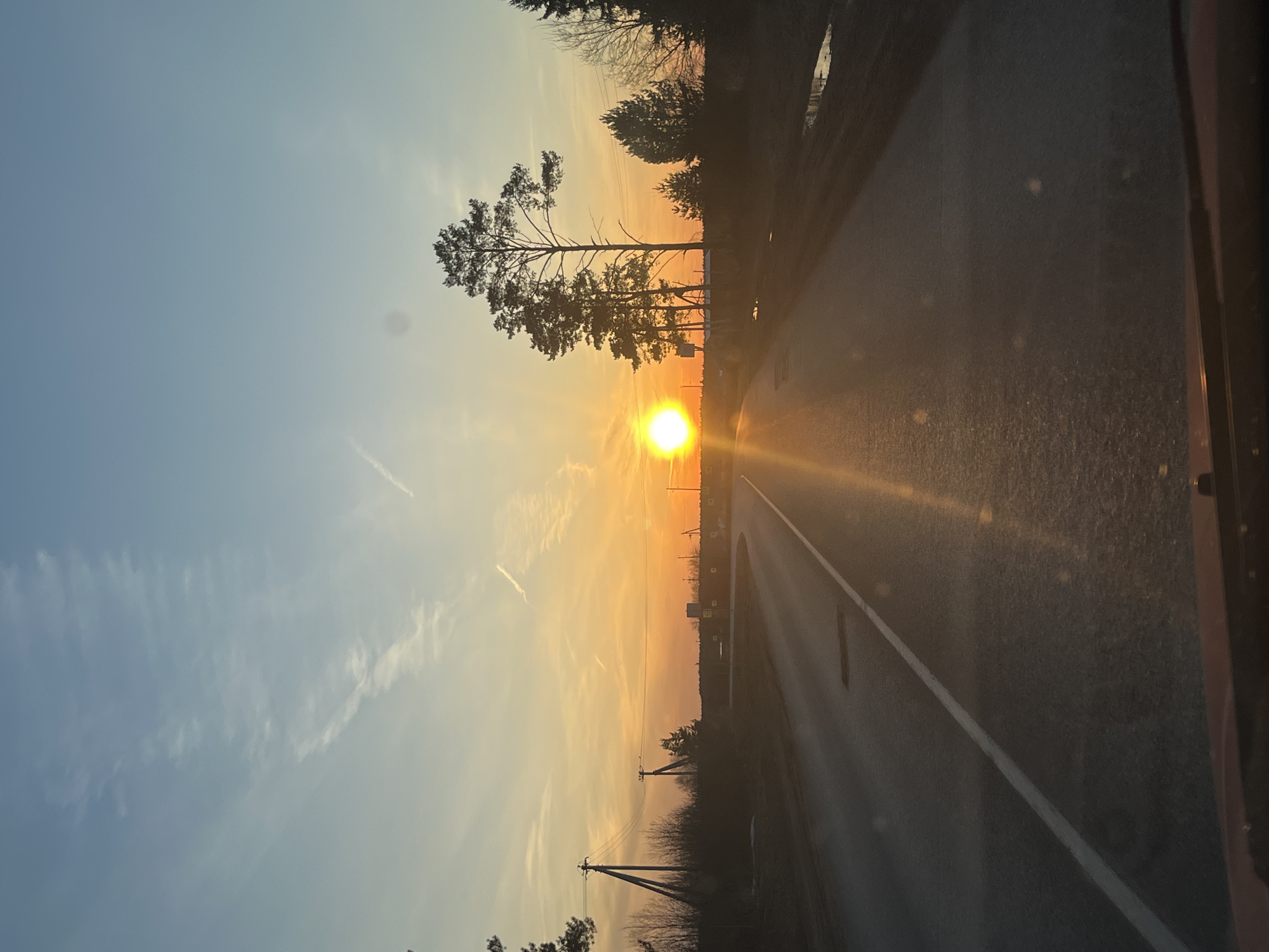 The image depicts a road stretching towards the horizon, presumably in Гаревая, Russia, as the user specified. The sun is setting directly ahead, casting a bright glare and long shadows along the road. Trees line the left side of the road, silhouetted against the vibrant sunset sky. Power lines and poles are also visible on the left. The sky transitions from a light blue at the top to orange and yellow near the horizon. The road itself is asphalt with a single white line marking the lanes. The image appears to have been taken from within a vehicle, based on the internal car panel that is seen. The time is likely late afternoon or early evening, given the position of the sun.