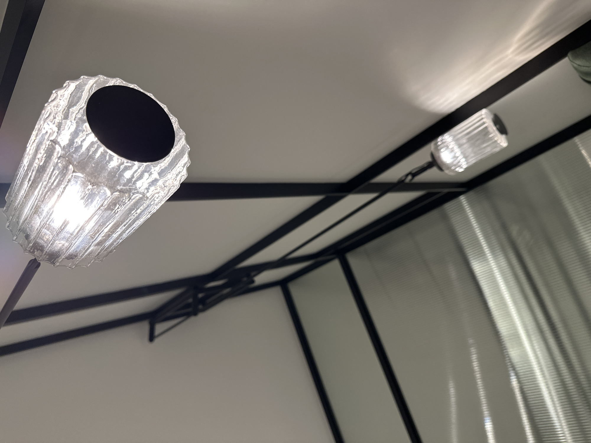 The image shows an interior space, likely under a gazebo or similar structure. The structure is defined by a black metal frame against white walls. Two light fixtures are visible. One is cylindrical, clear, ribbed glass with a dark, possibly black, interior opening, appearing to be illuminated from within. The second fixture is smaller, also clear glass, and similarly illuminated. Light reflects off the ceiling, creating bright patterns. A translucent curtain or panel is visible in the background. Based on the image, it is impossible to definitively identify the location as Tbilisi, Georgia.