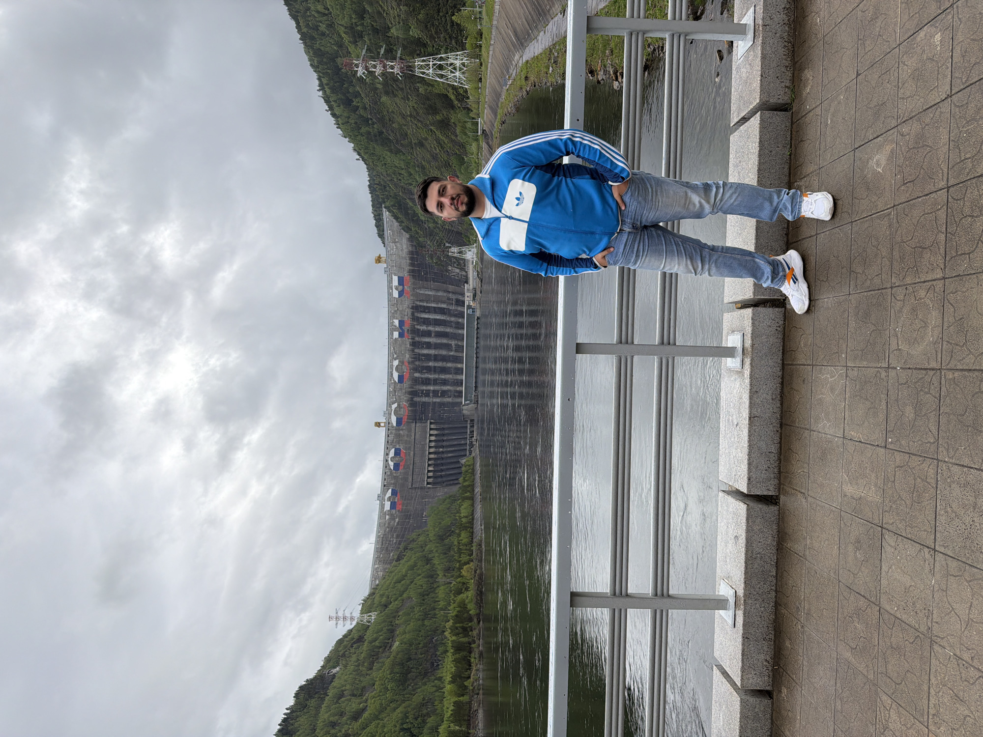 The image depicts a man standing on a paved platform, likely a walkway, adjacent to a railing. Behind the railing is a body of water, and in the background is a large dam structure adorned with what appear to be the colors of the Russian flag painted on its face. The surrounding environment includes green, forested hills. High voltage power transmission towers are visible on the left, ascending the hills. The sky is overcast with heavy cloud cover. The man is wearing a blue and white Adidas-branded tracksuit and white sneakers with orange accents. The location is the Sayano–Shushenskaya Dam on the Yenisey River, located near the city of Cheremushki, Russia.