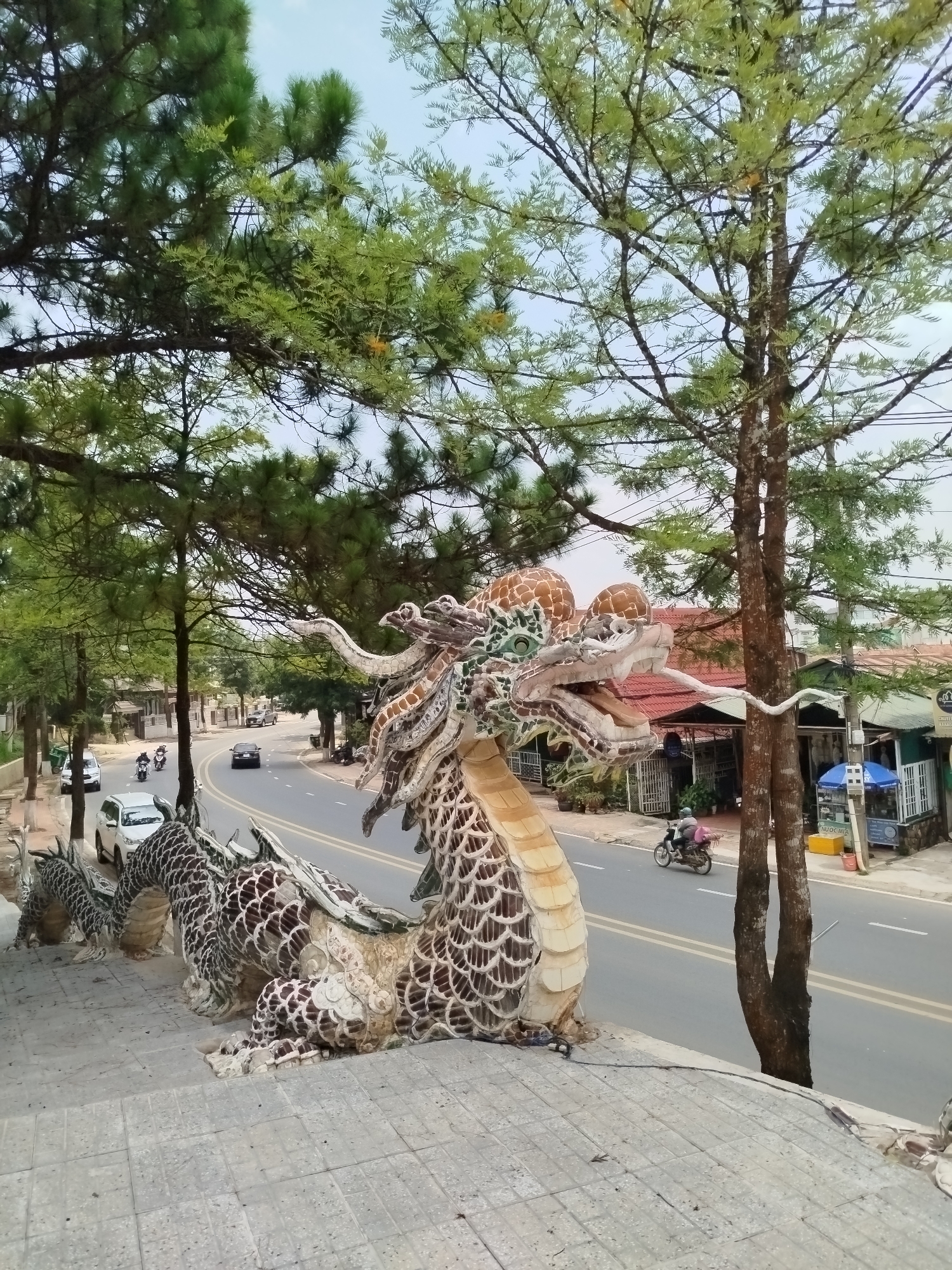 A full shot captures a lengthy mosaic dragon sculpture in Lang Biang - Đà Lạt, Vietnam. The dragon is composed of cream and brown tiles, with distinctive facial features and scale patterns. It's situated on a tiled terrace that overlooks a multi-lane street with vehicular traffic, including cars and motorcycles. Buildings and shops line the street beyond the traffic. Above the scene, trees with dense foliage frame the top part of the shot. The sky is overcast.