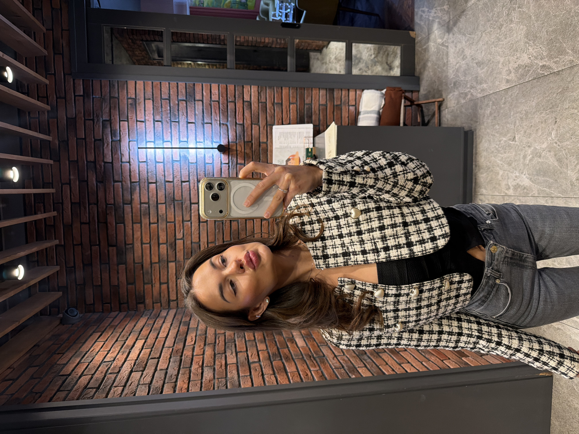 A woman with brown hair, wearing a black and white patterned jacket with pearl buttons and grey jeans, is taking a selfie with an iPhone. She has a light complexion and is making a kissing face. The background shows a brick wall with linear lighting and wooden shelves with spot lights on the left. To the right, there is a grey wall and a grey counter with some items on it. The scene appears to be indoors, possibly in a commercial space or residential area. The location is Krasnoyarsk, Russia, according to information provided.