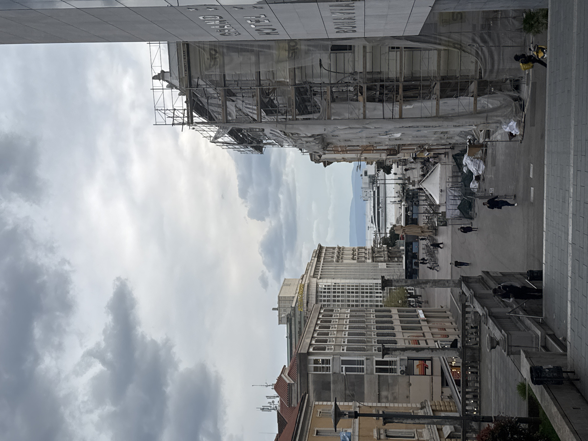 The image shows a view of the city of Rijeka, Croatia, on a cloudy day. The perspective appears to be from an elevated position. In the foreground and center, there's a street with several pedestrians walking in both directions. A building to the left is under construction, covered with scaffolding and protective wrapping. The structure has signage in Croatian. In the background, there's a glimpse of the Adriatic Sea, possibly the Kvarner Gulf. The buildings in the middle ground display varied architectural styles.