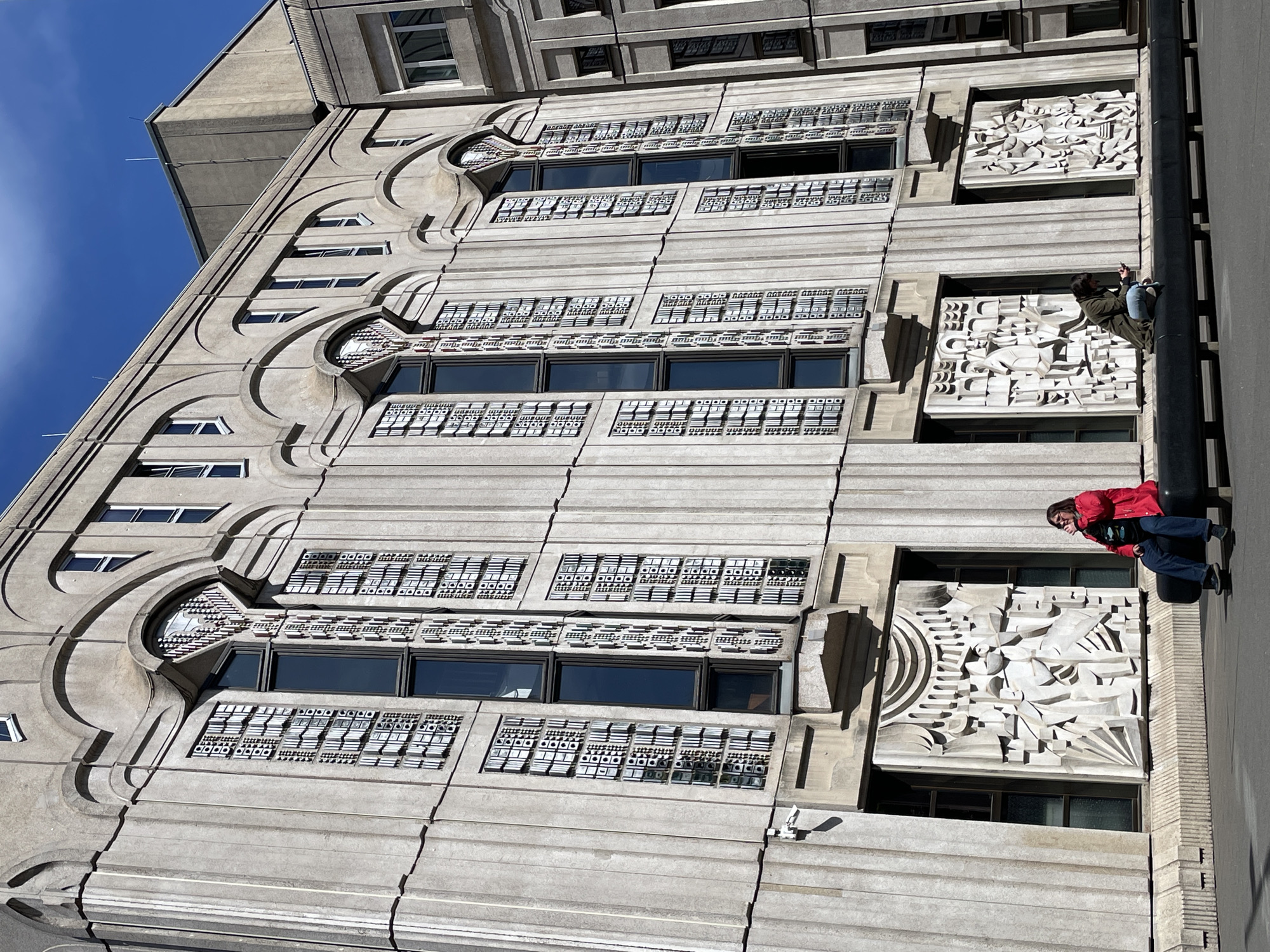 A photograph taken in Berlin, Germany, showcases the ornate exterior of a tall, light-colored concrete or stone building, likely from an Art Deco or early modern architectural period, under a clear blue sky. The image captures the building from a low-angle perspective, looking upwards at its intricate facade.
The building's design features prominent architectural details, including large, curved structural elements that frame sets of rectangular windows on the left side. Below and above many of these windows are decorative panels or grilles with repetitive, geometric, square-based patterns.
On the right side of the building, two large bas-relief sculptures are integrated into the facade, positioned between different floor levels. These sculptures display abstract, dynamic forms that suggest themes, possibly industrial or symbolic.
Two individuals are visible on the far right side of the building, leaning against a dark, vertical pipe or column that runs alongside the facade. The person closer to the bottom, wearing a red jacket, blue jeans, and glasses, appears to be observing the surroundings or taking a break. The second person, positioned higher up in a dark jacket, is also leaning against the same column.
The bright sunlight indicates daytime, casting strong shadows that accentuate the three-dimensional depth of the building's facade and sculptures. The sky is a vibrant, cloudless blue. No visible text is legible on the building's exterior.