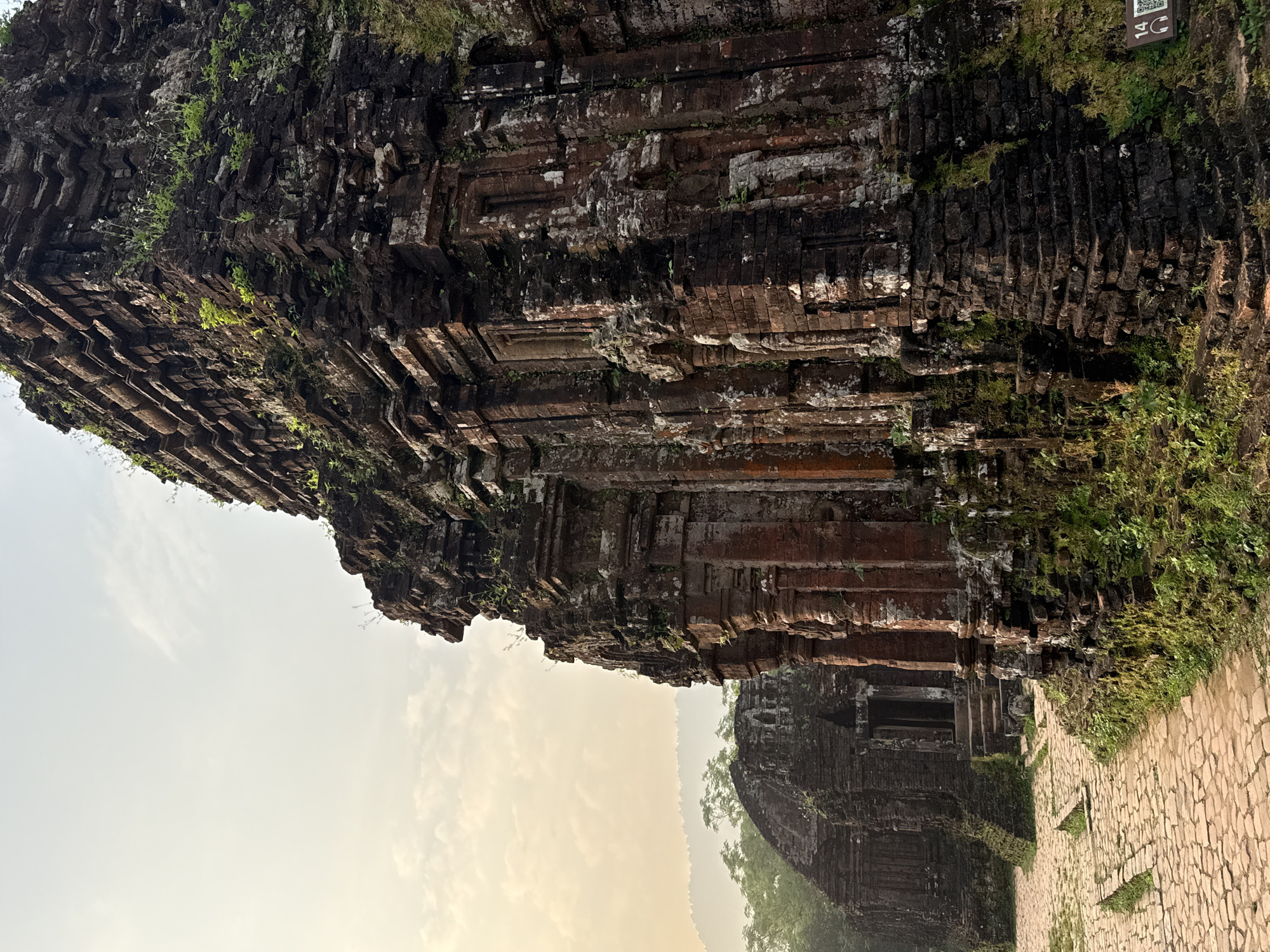 The image shows the ruins of the Mỹ Sơn temple complex in Đà Nẵng, Vietnam. The sandstone brick structures are decaying and covered with moss and vegetation. The sky is hazy, suggesting humid or foggy weather. A brick-paved pathway is visible alongside one of the structures.