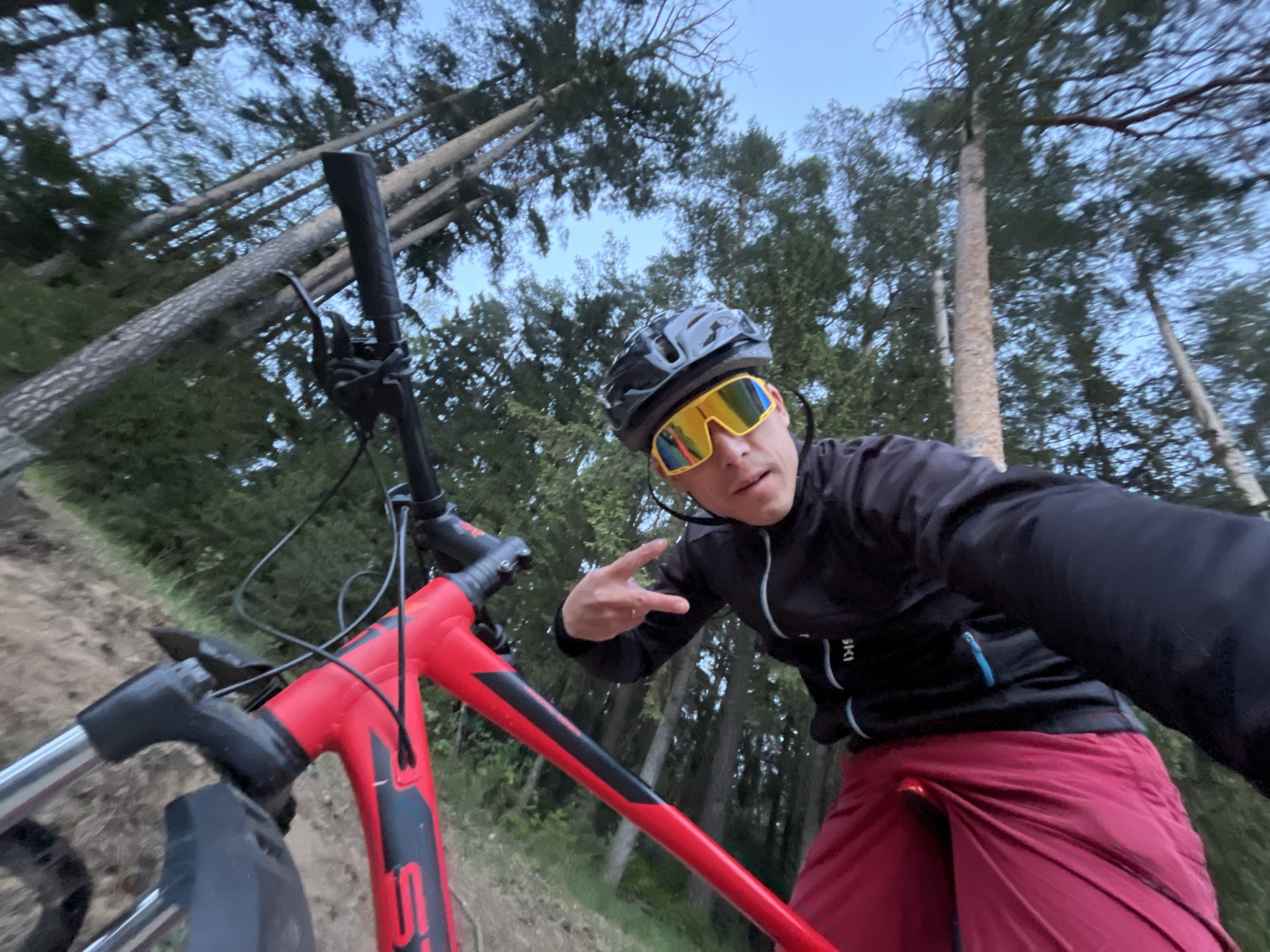 The image shows a man with short brown hair wearing a black helmet and yellow sunglasses, a black jacket, and red pants posing with a red mountain bike. He is making a peace sign with his right hand. The bike is partially visible in the foreground. The background consists of a forest setting with tall trees and a sky with a muted blue hue. The location is Chaikovsky, Russia.