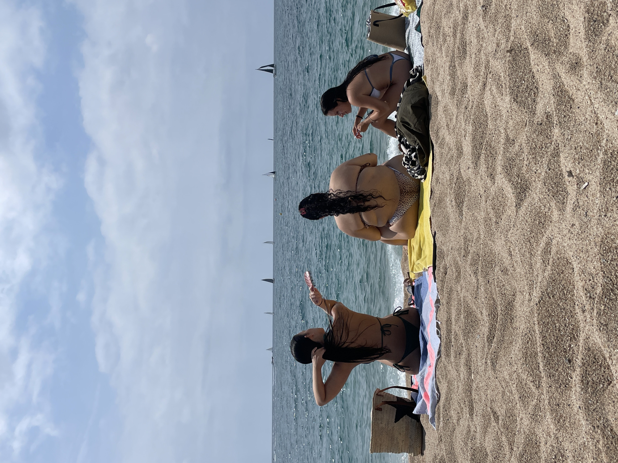 Three individuals are sitting on the sand at the beach in Arenys de Mar, Spain, facing the sea. The person on the left, viewed from behind, has long dark hair and is wearing a dark bikini. They are holding a comb and appear to be brushing their hair. In the center, another person with dark hair, also viewed from behind, is wearing a bikini with a leopard print bottom and a dark top. The person on the right, facing the center individual, has dark hair and is wearing a light-colored bikini. They are sitting on a light blue towel. In front of the individuals are beach towels and bags. The sea is calm with several sailboats visible on the horizon under a cloudy sky. The foreground is composed of coarse sand.