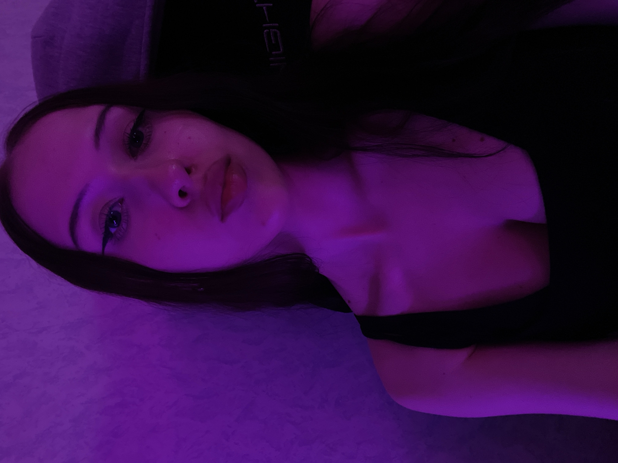 The image is a selfie of a young woman indoors, bathed in purple light. She has dark hair and is wearing a black tank top. The background appears to be a textured wall with a light fixture or a piece of furniture with the word "IGHT" visible. The woman is looking towards the camera with a neutral expression and her lips slightly parted. The scene is likely set in a residential or personal space.