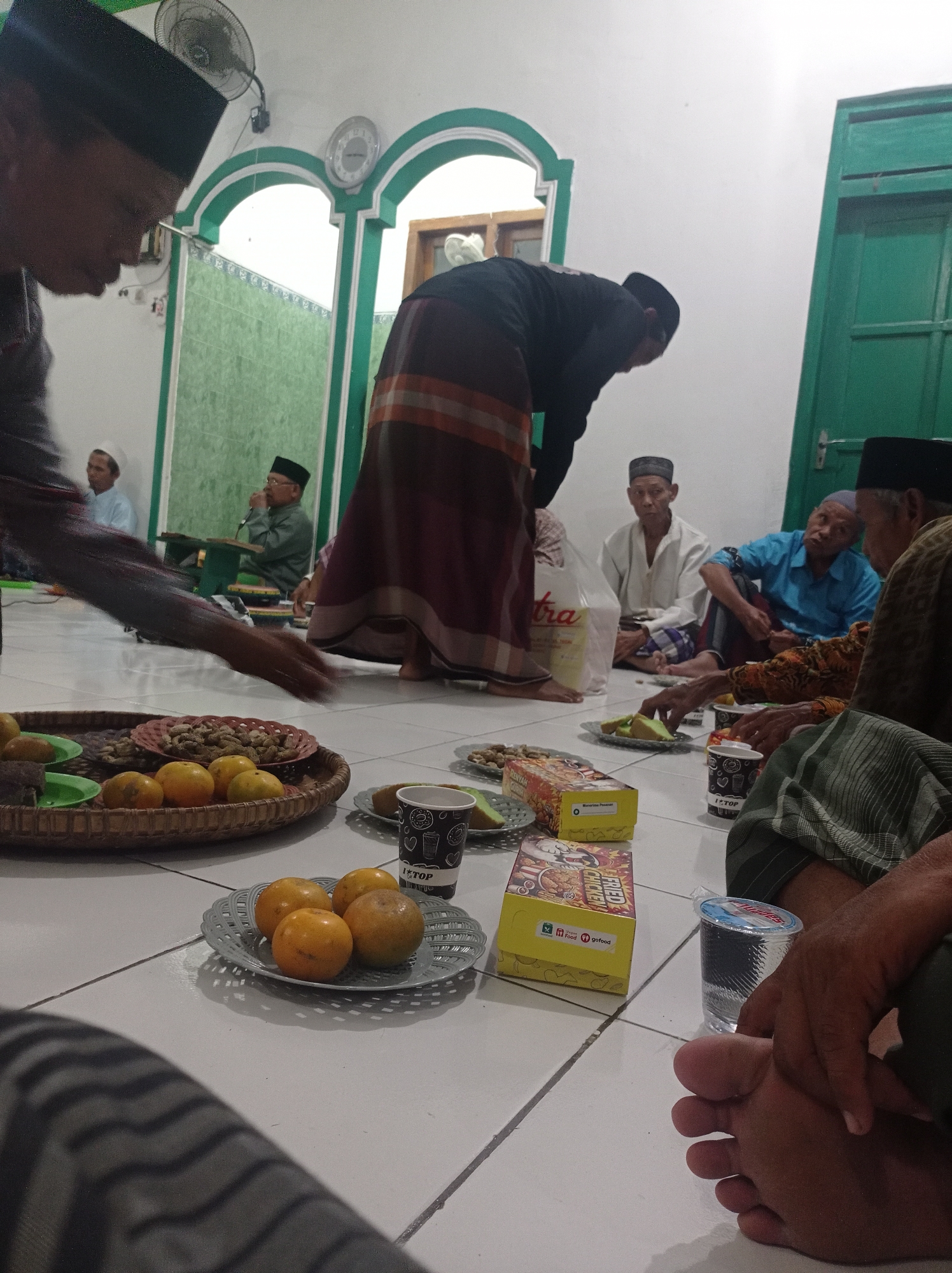 The image depicts a gathering of men, likely in a mosque or a similar community space, in Maribaya, Indonesia. Several men are seated on the floor, some engaged in conversation. One man in the foreground is in the process of serving food from a large platter. Food items, including oranges, peanuts, and what appears to be cake or pastries, are spread across a woven mat and on plates. Disposable cups and boxes of food, including fried chicken, are also visible. The environment is indoors, with a tiled floor and white walls. The presence of traditional Islamic attire, such as peci hats and sarongs, suggests a cultural or religious event. The overall scene conveys a sense of community and shared meal.