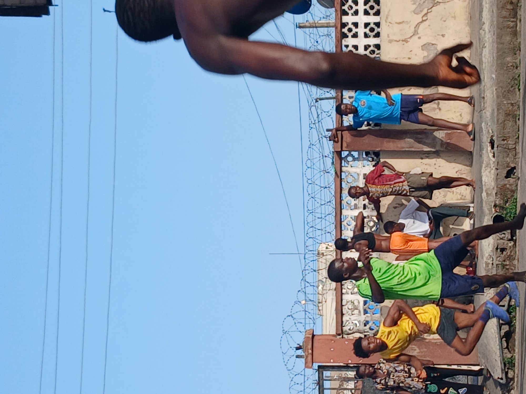 A medium-angle, outdoor shot depicts at least seven Black men, positioned along a concrete wall topped with razor wire, under a clear blue sky in Lagos, Nigeria. The men are engaged in some form of physical training or exercise.
The foreground features the partially visible upper body of a dark-skinned person, likely male. Behind this, a line of men stretches horizontally, most wearing casual athletic attire. One man is wearing a bright yellow shirt and gray shorts. Another is in a neon green shirt and dark blue shorts. Other men are in various other casual attire, including a blue shirt and another with a patterned shirt. The men appear to be performing some form of physical activity, possibly stretching or calisthenics. They seem to be in a line, facing either forward or sideways, with the building and razor wire behind them.
The concrete wall is topped with a significant amount of razor wire. The wall appears weathered and textured, with peeling paint. Behind the wall, there is a building with ornamental metalwork.
The sky is clear blue, devoid of clouds. Electrical wires are visible across the sky, enhancing the urban backdrop.
The lighting is bright and sunny.
