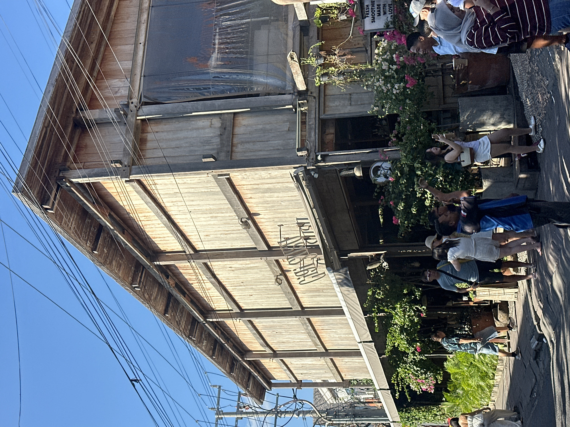 This is a photograph of a street scene in North Kuta, Indonesia. The focal point of the image is a multi-story wooden building with a sloped roof and exposed wooden beams. The building has a rustic appearance, with weathered wood siding. In front of the building, there is a covered awning, and lush green plants and flowering bushes are visible. A sign with the words "FRESH SMOOTHIES MADE WITH LOVE" is also present.
Several people are in the foreground and on the sidewalk in front of the building. They appear to be a mix of locals and tourists, dressed in casual clothing. Some individuals are walking, while others are standing and conversing. One woman is holding a smartphone, possibly taking a picture. The street is paved with cobblestones in some areas and asphalt in others. Power lines are strung across the top of the image against a clear blue sky. The overall impression is a lively, outdoor commercial area.