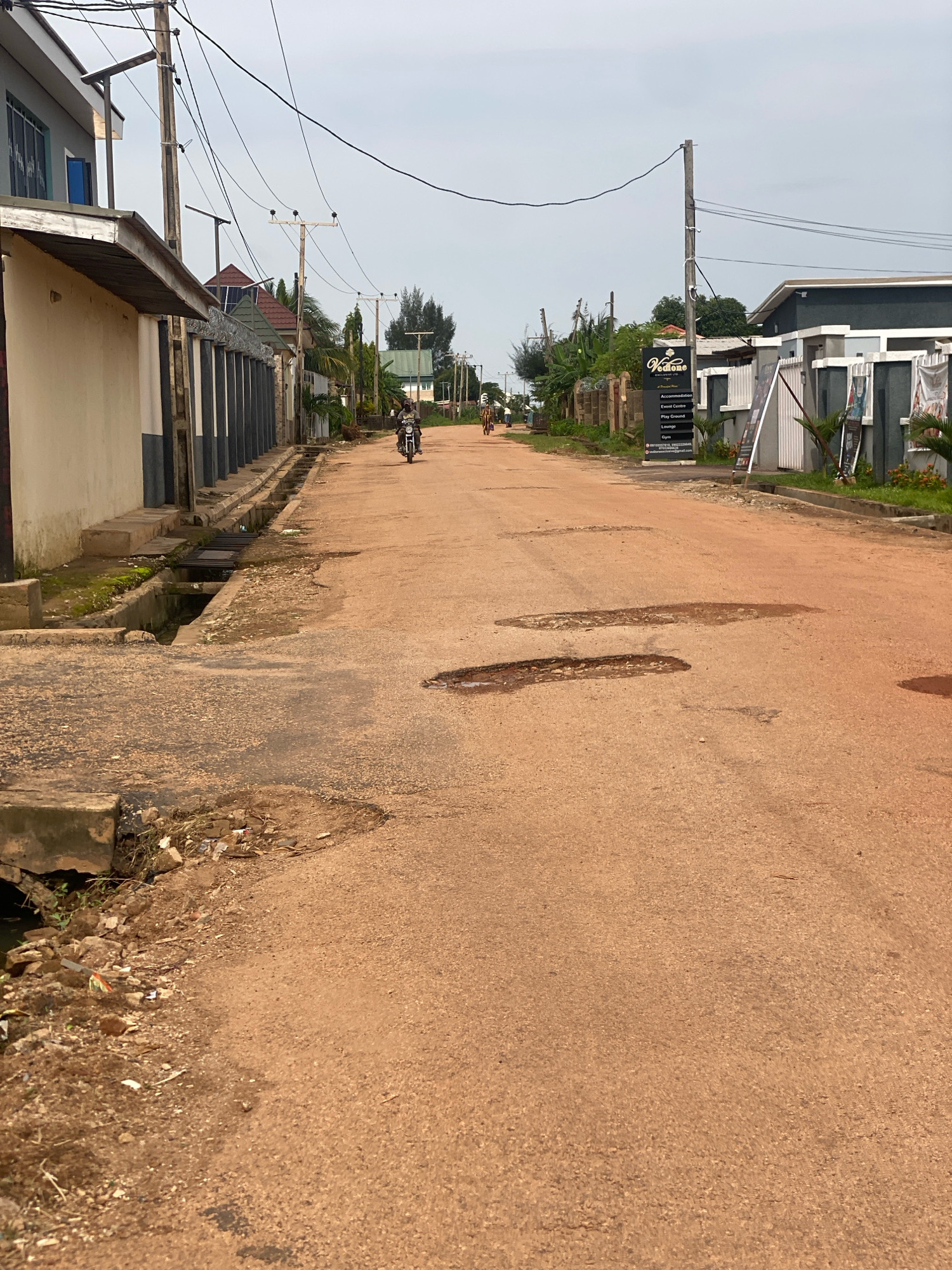A street in Barden Yana, Nigeria is shown in this eye-level, medium shot. The street is paved with reddish-brown asphalt and has visible potholes. On the left side of the street is a row of buildings with a drain running along the front. One building is light beige with dark grey accents, and another has a white facade with dark grey vertical pillars. The right side of the street features a modern building with a grey facade, white fences, and large glass windows. A black sign for "Vedonia" with text listing "Accommodation, Event Centre, Play Ground, Lounge, Gym" is posted near the building, along with a white A-frame sign. Overhead, a network of utility poles and wires crisscrosses the sky, which is overcast. Two people are visible on motorcycles on the street, moving away from the viewer. The overall scene depicts a somewhat neglected but functional urban environment.