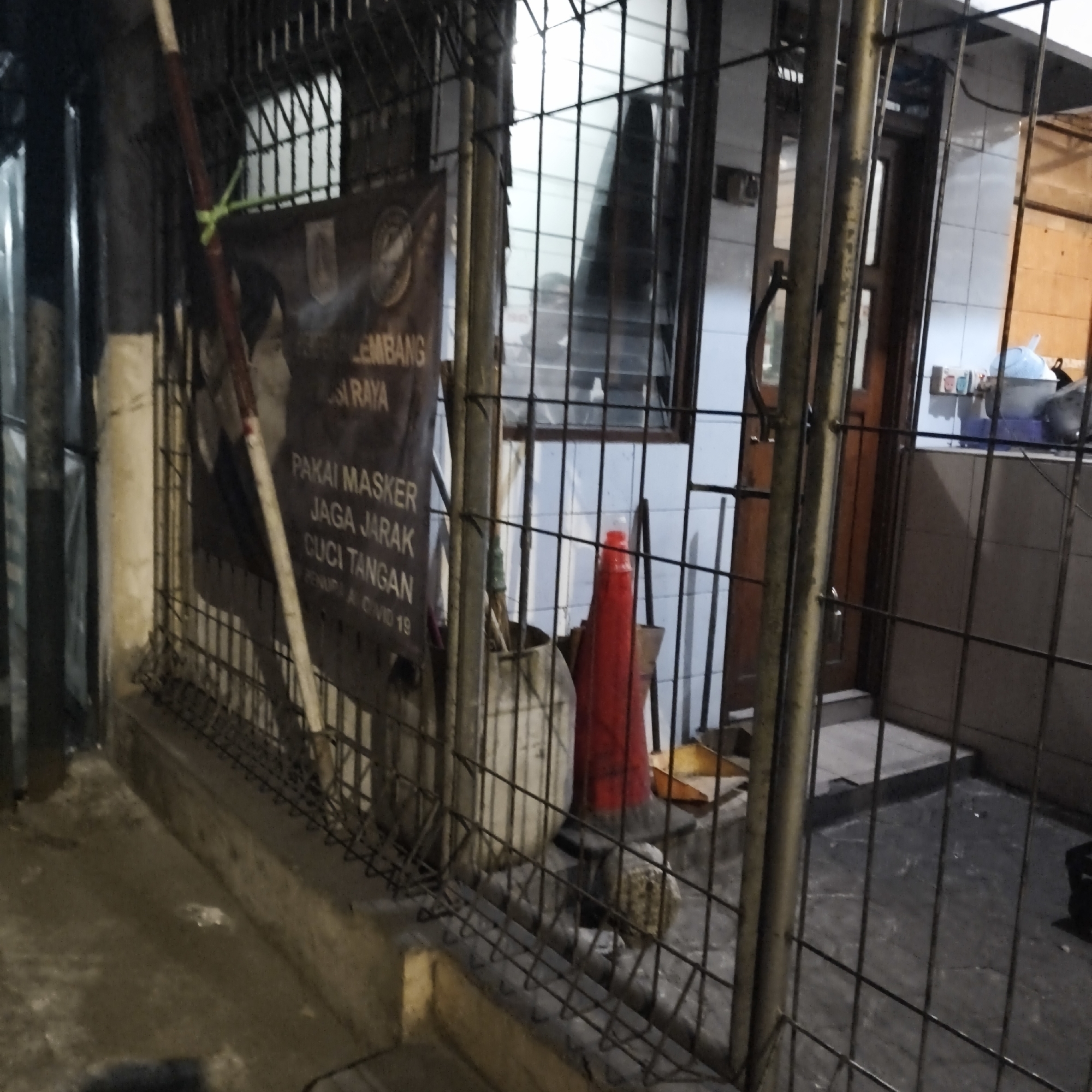 The image depicts a nighttime scene outside a building with a metal fence in the foreground. The fence encloses a narrow walkway or porch area. Behind the fence, a dark banner with Indonesian text is visible, hanging from a pole. The text on the banner appears to be related to health protocols, mentioning "PAKAI MASKER" (wear a mask), "JAGA JARAK" (keep distance), and "CUCI TANGAN" (wash hands), along with "COVID 19". To the right of the banner, a red traffic cone stands near a large, light-colored cylindrical object, possibly a trash can or storage container. Further to the right, a wooden door with glass panels leads into the building. The building exterior features tiled walls. On a shelf inside the building, several items are visible, including what appears to be a light blue lid of a container and possibly some bottles. The ground outside is dark and wet, suggesting it might be raining or recently rained. The overall impression is a utilitarian outdoor area, possibly at the entrance to a shop or residence, with a clear emphasis on public health messaging. The location is identified as South Jakarta, Indonesia.
