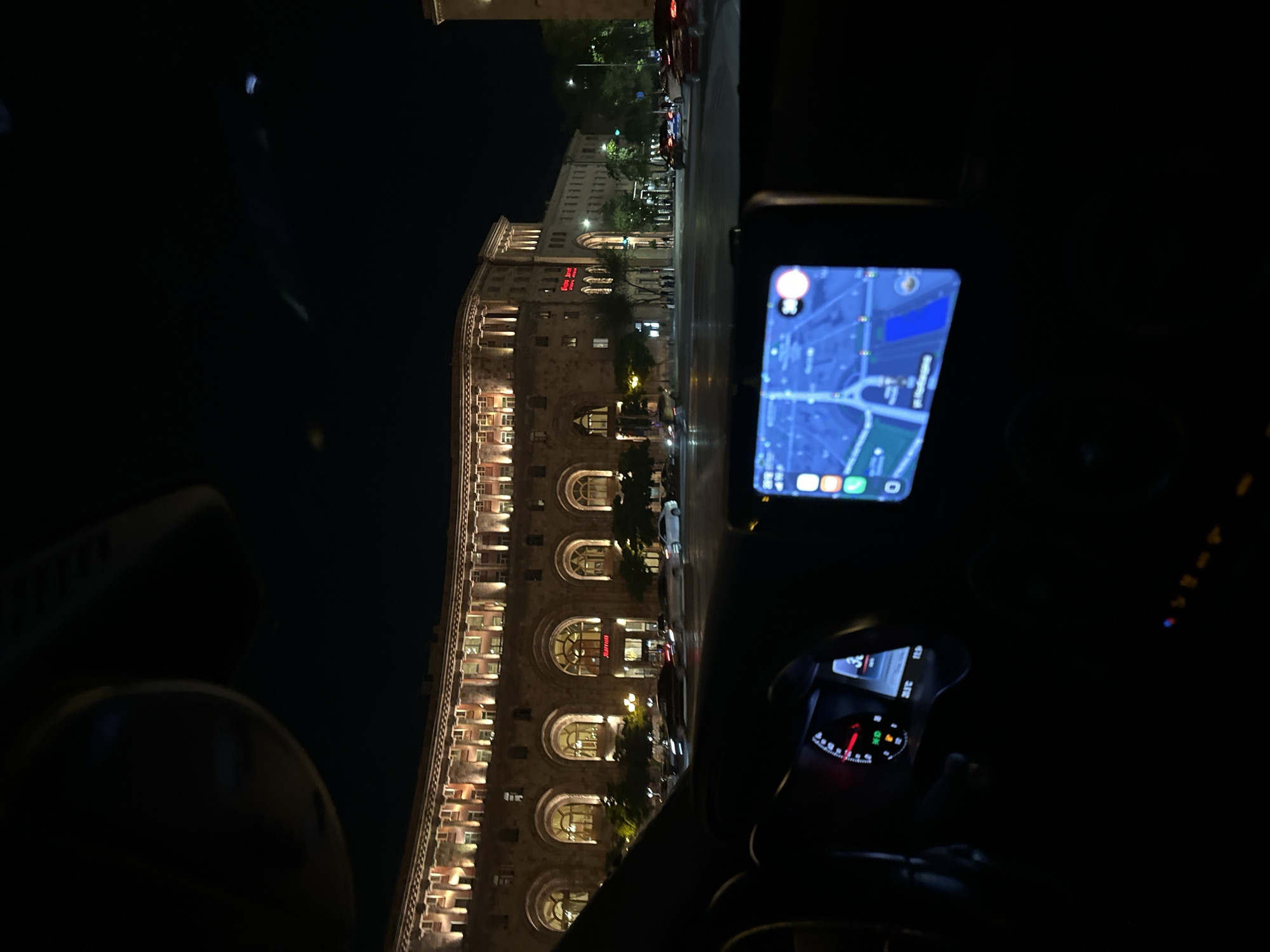 The image shows a nighttime view from inside a car in Yerevan, Armenia. The car's dashboard is visible in the foreground, with a navigation screen displaying a map. The main subject of the image is a large, ornate building with illuminated windows, characteristic of Soviet-era architecture. This building is Republic Square, a prominent landmark in Yerevan. The square is bustling with activity, with cars and pedestrians present. The overall atmosphere is one of a vibrant city at night.