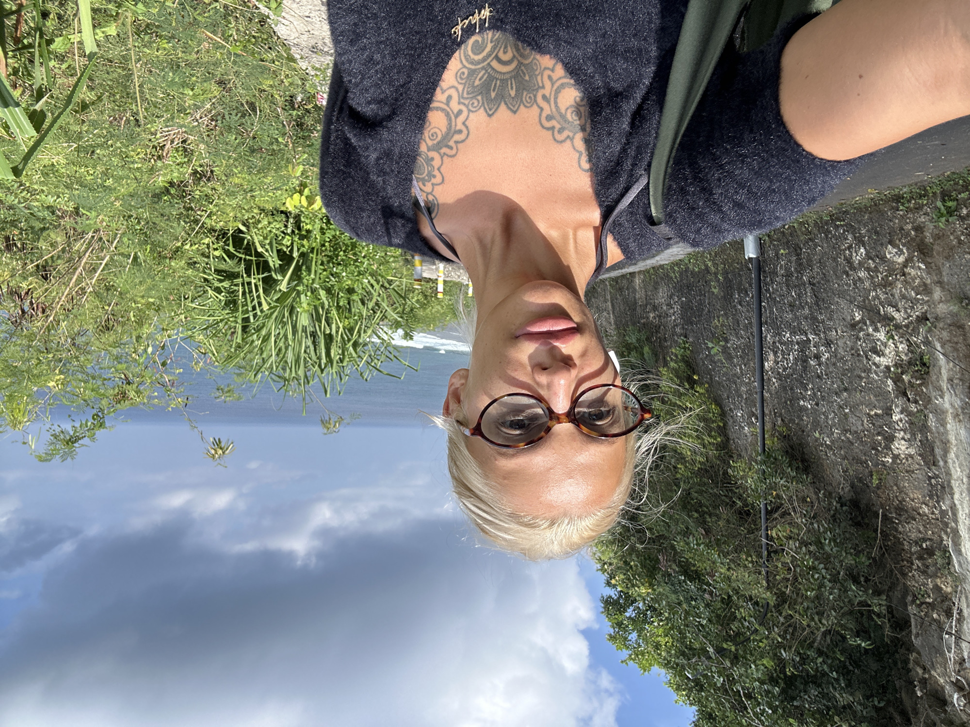 A blonde woman wearing glasses and a black sweater takes a selfie outdoors. She has a tattoo on her chest. Behind her is a rocky wall, a paved road, lush green vegetation, and the ocean under a cloudy sky. The location is the cliffside road leading to Nyang Nyang Beach in Kuta Selatan, Bali, Indonesia.
