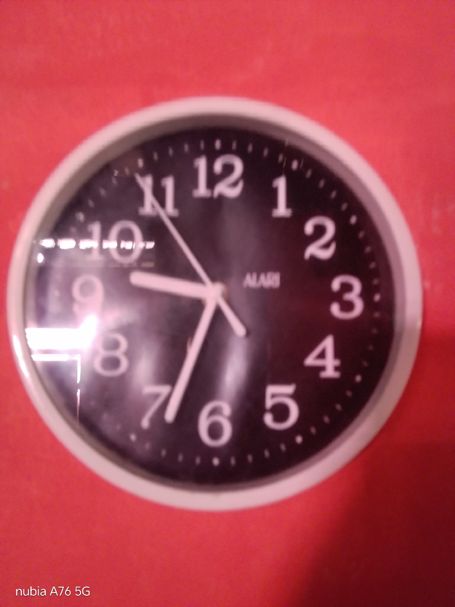 A close-up, slightly blurry image shows a black analog clock with white numbers and hands against a red background. The clock face has a white bezel and is slightly tilted. The numbers visible are 1, 2, 3, 4, 5, 6, 7, 8, 9, 10, 11, and 12. The clock hands are white and appear to indicate a time slightly after 9:10. The brand name "ALARI" is visible on the clock face. The lower-left corner of the image displays the text "nubia A76 5G". The red background appears to be a wall with a textured surface, possibly painted. There is a bright reflection on the clock face, obscuring some of the details. The image does not contain any specific geographical markers or contextual information that would allow for its placement within the city of Cakung, Indonesia.