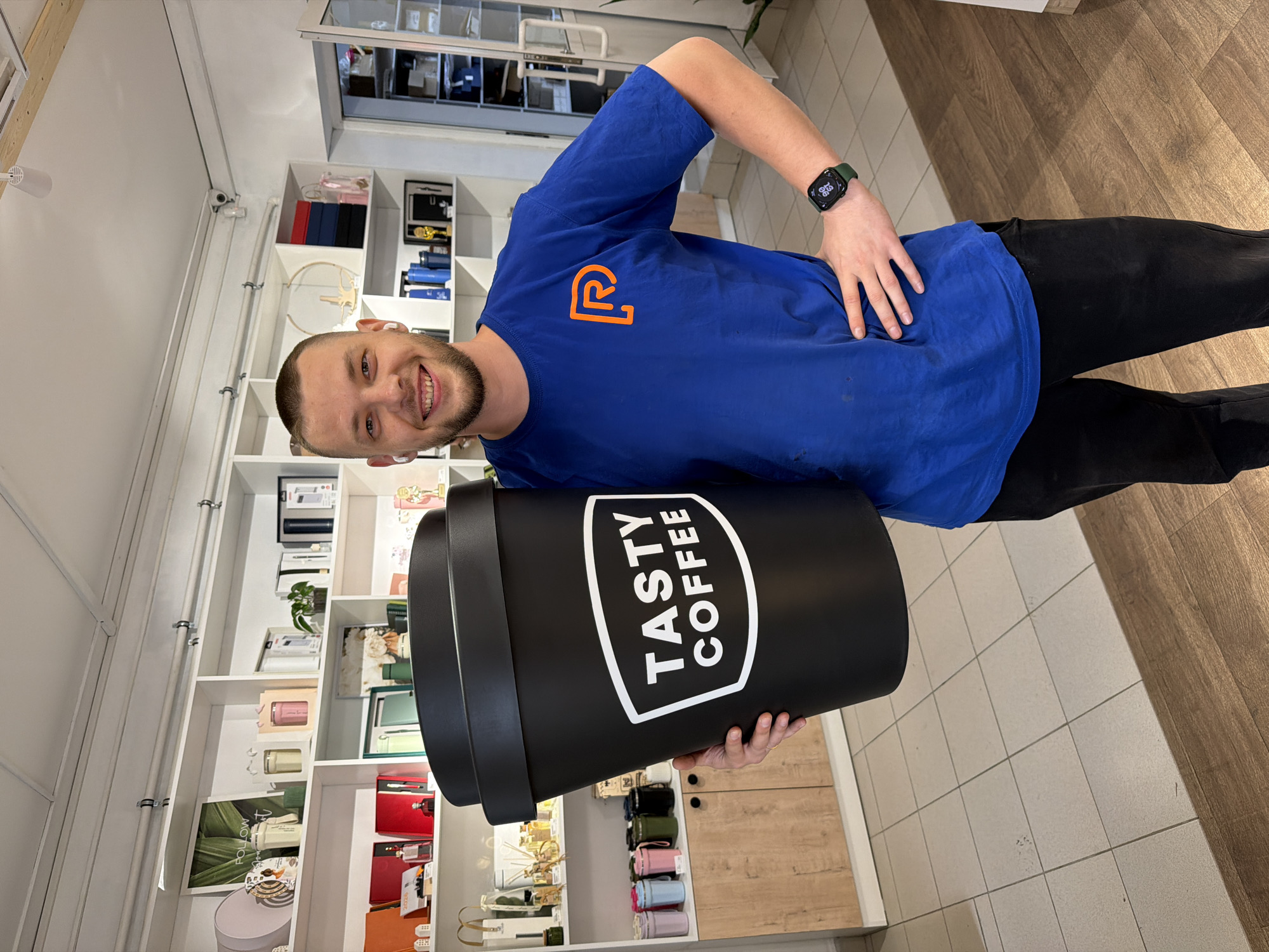 A Caucasian male stands smiling and holding a large black coffee cup with the words "TASTY COFFEE" printed on it. He is wearing a blue T-shirt with an orange logo on the left chest, black pants, and a smartwatch on his left wrist. He has short brown hair and a beard. He is standing inside a store or showroom, with display shelves filled with various items in the background. The floor is tiled with a mix of white and wood-patterned tiles. The location is Chaikovsky, Russia.