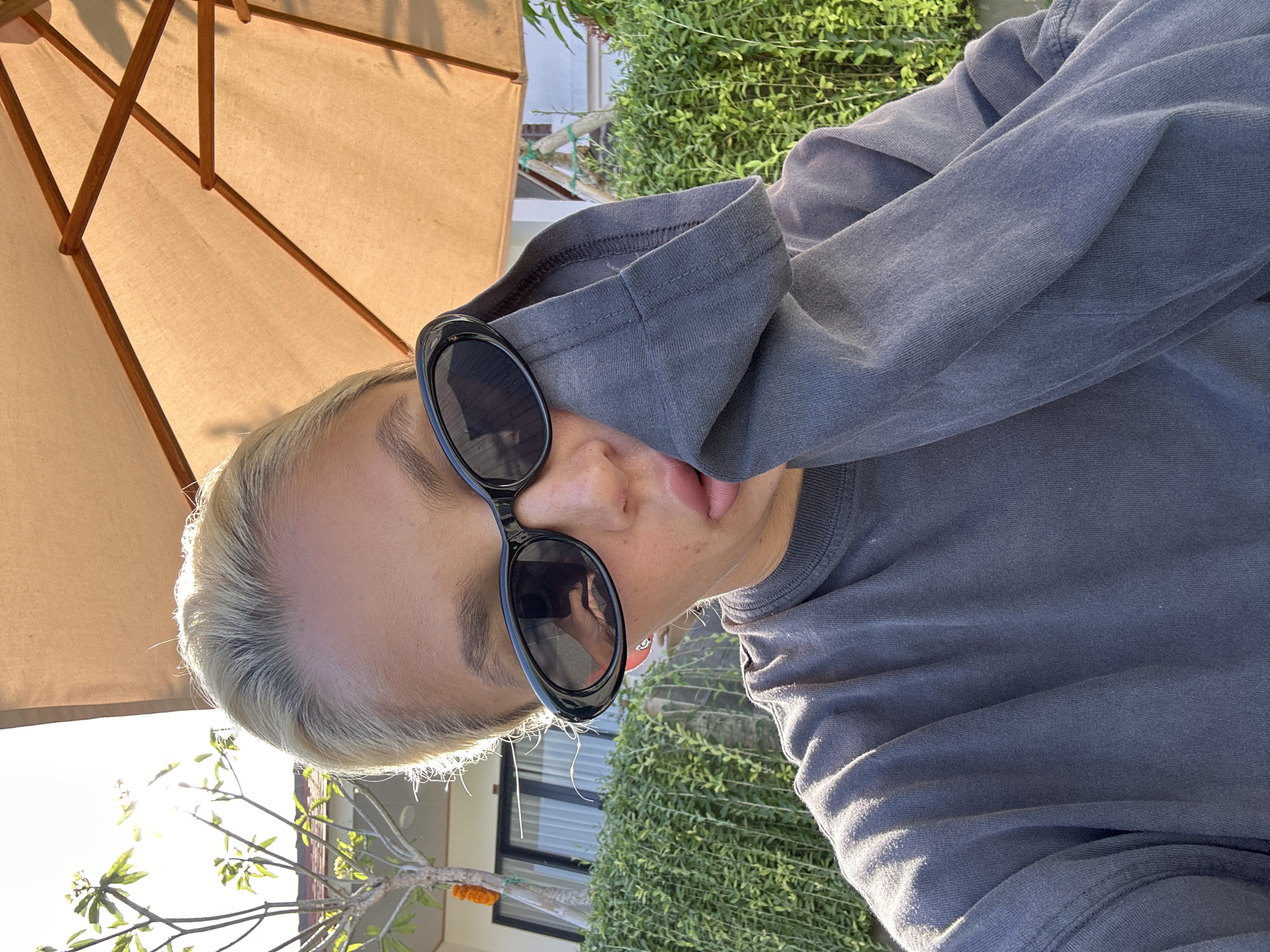 A woman with blonde hair pulled back and wearing black sunglasses and a grey long-sleeved shirt is captured in a selfie. She is holding the sleeve of her shirt up to her face, obscuring her mouth and chin. The background includes lush green foliage, a building with windows, and a large, light-brown umbrella casting shadows. The lighting suggests it is daytime, with the sun creating a slight glare. The scene appears to be an outdoor area, possibly a resort or a home, in Kuta Selatan, Indonesia, given the tropical vegetation and architecture.