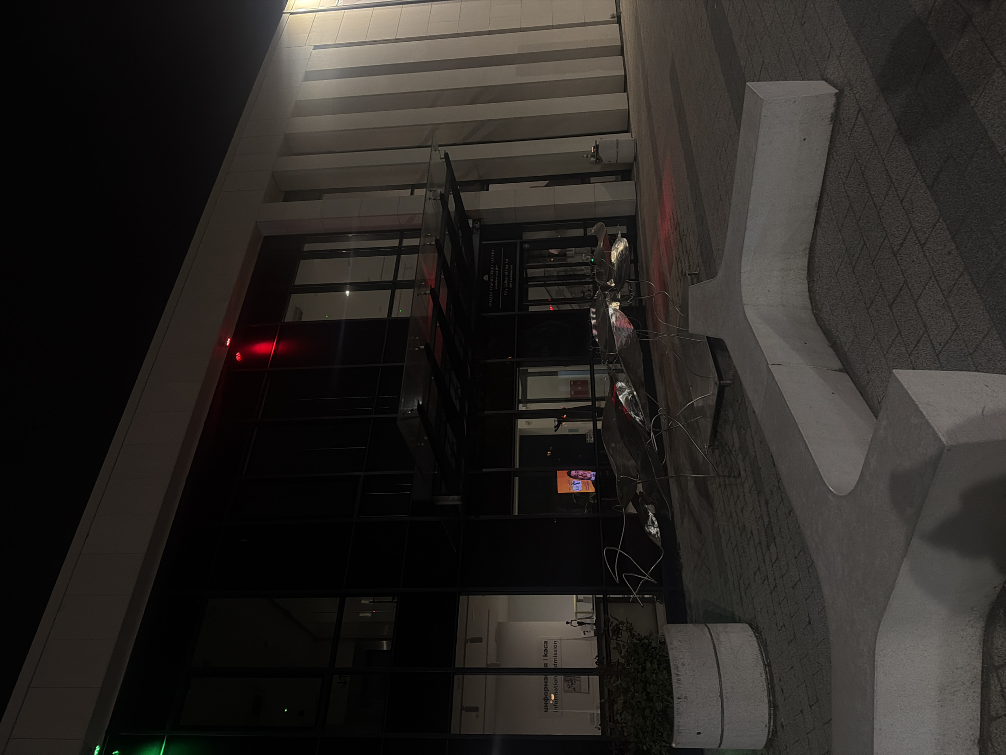 The image depicts a modern building in Plovdiv, Bulgaria, captured at night. The multi-story structure features a sleek design with a prominent glass facade and light-colored horizontal bands along its upper sections. Artificial lighting, including red and green lights, illuminates parts of the building and its surroundings against the dark night sky.
In the foreground, a large, light-grey concrete structure shaped like a stylized letter 'Y' or an abstract form is positioned on a paved area made of textured grey bricks. Adjacent to this, a smaller cylindrical concrete planter containing some foliage is visible. Metallic, abstract, wire-like elements, possibly part of a sculpture or lighting installation, are also present near the concrete 'Y'.
Through the building's large glass windows, parts of the interior are visible. One window shows a small screen displaying a person's face. Another window reveals an interior sign with text: "информация | каса" (information | cash desk/ticket office) and "Information | admission". A prominent black vertical sign mounted on the building's exterior displays the name "ПРАЗКА ХУДОЖЕСТВЕНА ГАЛЕРИЯ" (Prazka Art Gallery), along with "City Gallery of Fine Art" and "Founded since 1881", identifying the building as an art gallery.
The scene is quiet, with no people or explicit activities taking place, emphasizing the architectural details and the artificial illumination of the urban environment after dark.