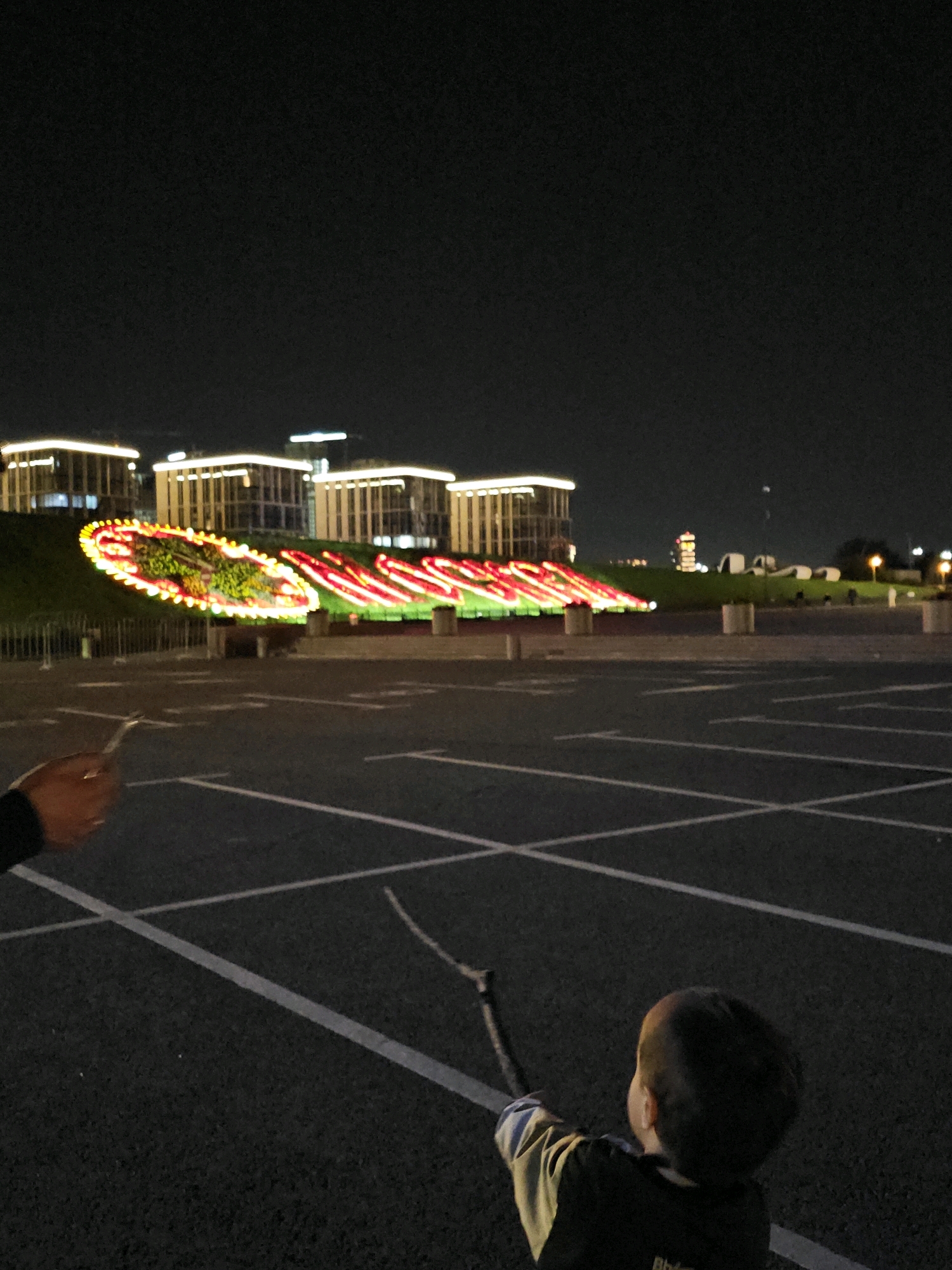 An outdoor, eye-level, long shot captures a dimly lit, night scene of a parking lot with a young child in the foreground and a brightly lit floral arrangement in the background. The child, facing away from the camera, is holding a stick. A hand holding a small object is visible on the left side of the frame. In the distance, a landscaped area features a large, oval-shaped floral display with lights in red and yellow. Behind the floral display are several modern, multi-story buildings illuminated with white lights. The parking lot is paved with asphalt and marked with white lines. The sky is dark. This scene is likely located in Moscow, Russia, near the VDNKh park, where such light displays are common during the holiday season.