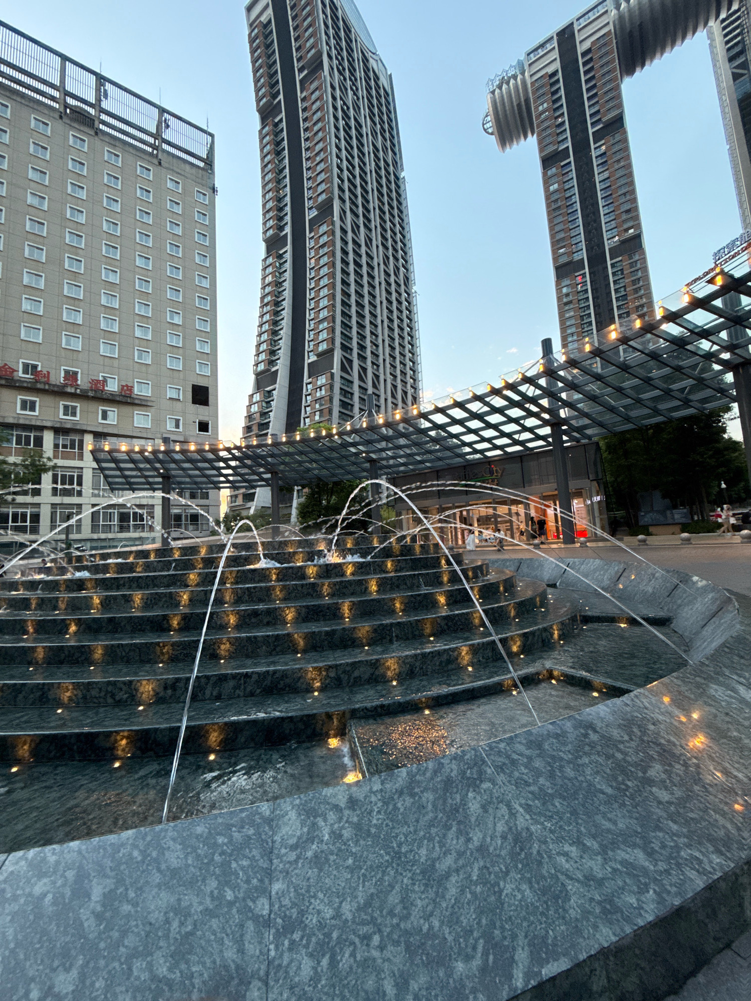 In the Yuzhong District of China, an outdoor tiered fountain with illuminated steps flows with water. Behind the fountain, a modern glass-covered walkway with decorative lights spans across the foreground. Beyond the walkway, several tall, contemporary buildings rise into the sky. To the left, a lighter-colored, rectangular building with numerous windows stands. To the right and center, taller, darker buildings with intricate architectural designs are visible. The sky above is clear and pale blue, suggesting it is either early morning or late evening. A few people are visible walking on the pavement in the background.