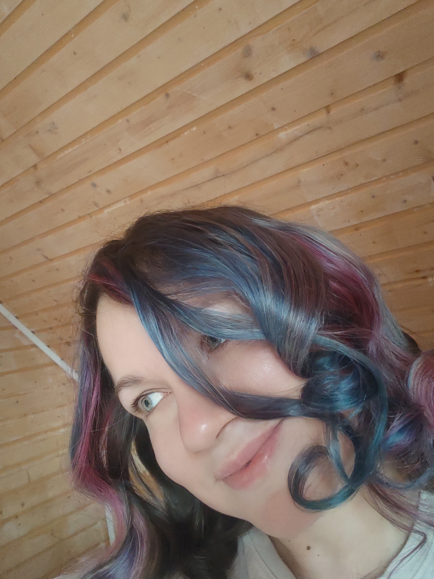A selfie is taken from a low angle, looking up towards the subject's face. The subject has shoulder-length, curly hair that is dyed with a mix of blue and pink hues. A strand of hair partially covers the right side of her face, obscuring her eye. She has fair skin and light-colored eyes that are partially visible. She is smiling softly. In the background is a wooden ceiling. Given the location of Свистуха (кузяевский С/о), Russia, the interior suggests a home or cottage.