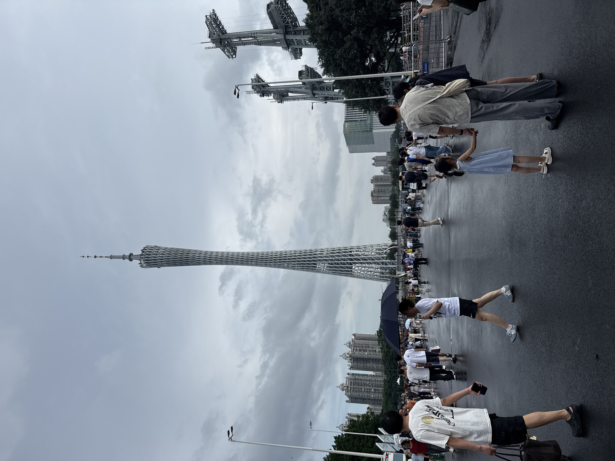 In Tianhe District, China, a wide, wet, paved area is filled with pedestrians under a cloudy, overcast sky. Dominating the background is the Canton Tower, a tall, slender lattice tower with a distinctive twisted design, reaching towards the sky. To the right of the Canton Tower, two large, industrial-looking metal structures, possibly part of a construction project or other infrastructure, are visible. The midground features several multi-story buildings, some with ornate architectural details, and trees lining the street.
The foreground is populated by people of various ages walking in different directions. A man in a white t-shirt and black shorts walks towards the viewer, holding a dark bag in his left hand and a phone in his right. To his right, a woman in a white t-shirt and black shorts walks away from the viewer, holding a dark object. Further ahead, a man in a beige shirt and grey pants holds hands with a young girl in a light blue dress. Several other individuals and groups are scattered throughout the scene, some with umbrellas, indicating recent rain or the possibility of precipitation. The wet pavement reflects the overcast sky and the surrounding structures.