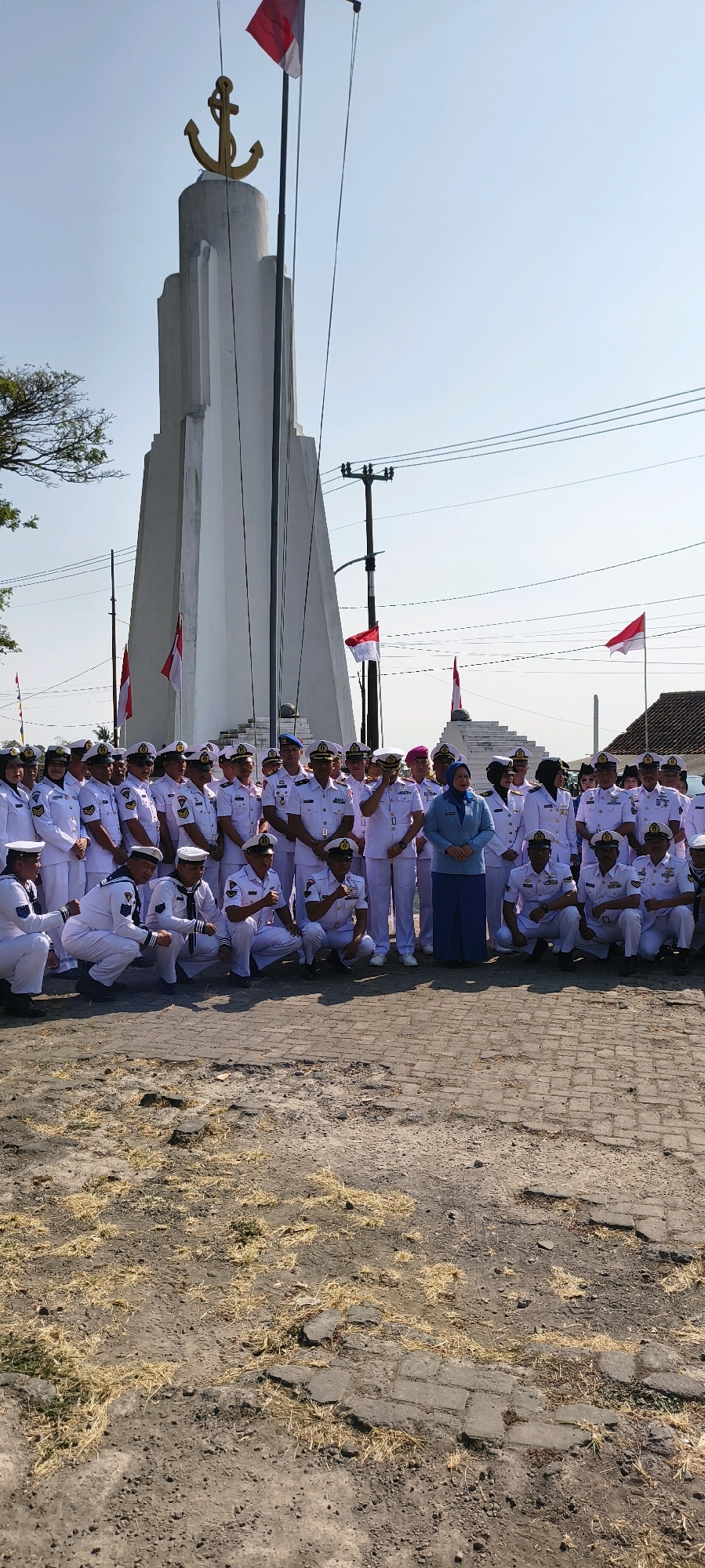 A group of people, predominantly men in white uniforms with gold accents, stand and kneel in front of a tall, white monument topped with a golden anchor. Several red and white flags are visible, some flying from poles and others attached to the monument. A woman in a blue jacket and pants stands in the center of the group. The environment appears to be outdoors, with a clear blue sky and some trees in the background. The ground is rough, with patches of dirt, grass, and cobblestone. The scene suggests a formal gathering or ceremony. The image was taken in Sadamantra, Indonesia.