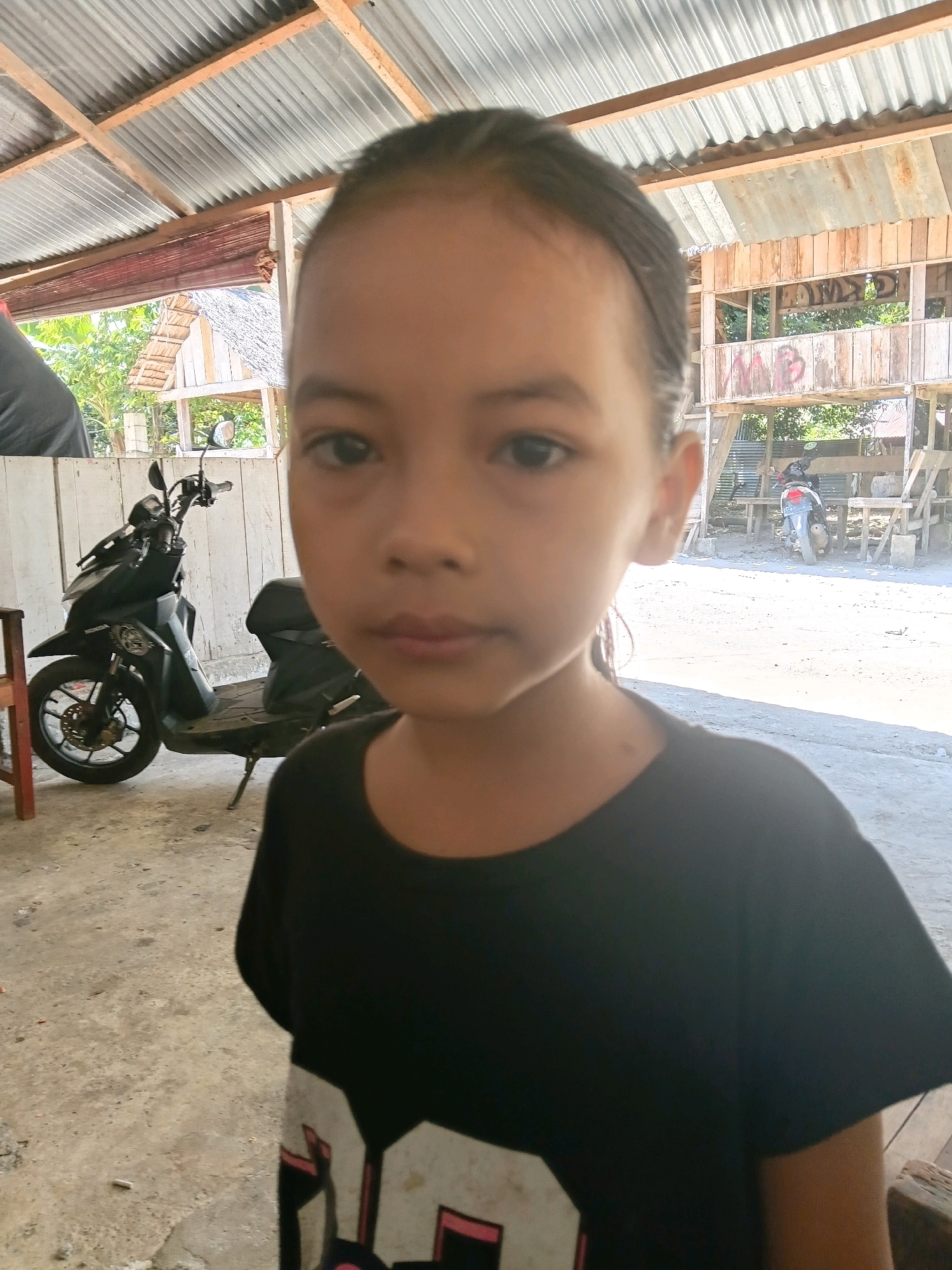 In Cotmanyang, Indonesia, a young girl with dark hair pulled back stands in a close-up shot. She is wearing a black t-shirt with white and pink graphics on the front. Her expression is neutral as she looks directly at the camera. In the background, a black scooter is parked. Behind the scooter, a rustic wooden structure with a corrugated metal roof is visible, suggesting an outdoor, possibly rural or informal setting. The ground appears to be a dirt or concrete surface.