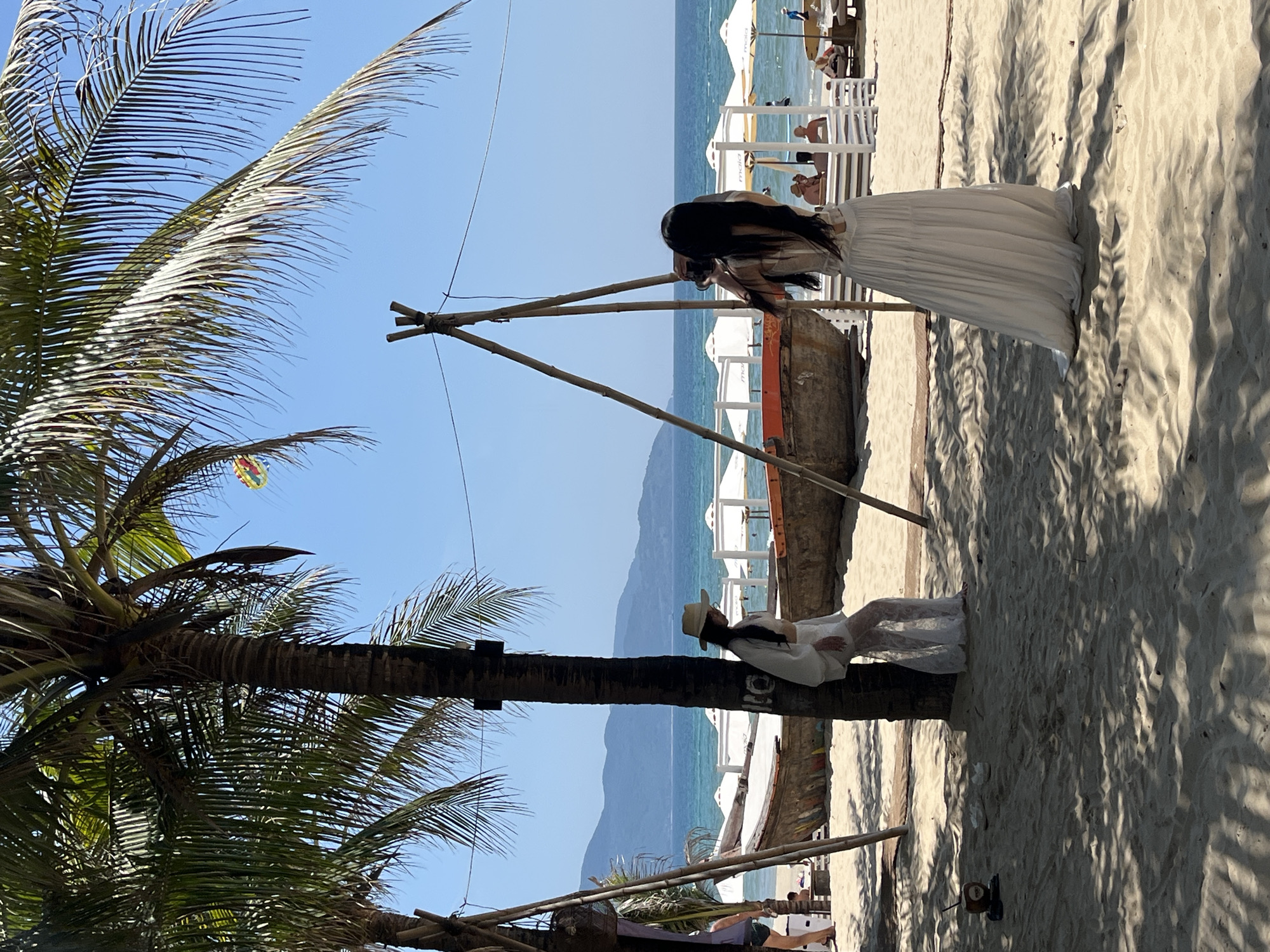 A beach scene in Đà Nẵng, Vietnam, features two women. One is in a long white dress, photographing the other, who is seated on a palm tree trunk wearing a white dress and hat. The background shows white umbrellas, the ocean, and distant mountains. A wooden structure stands near the women. The sand is light colored and displays shadows from the palm trees.
