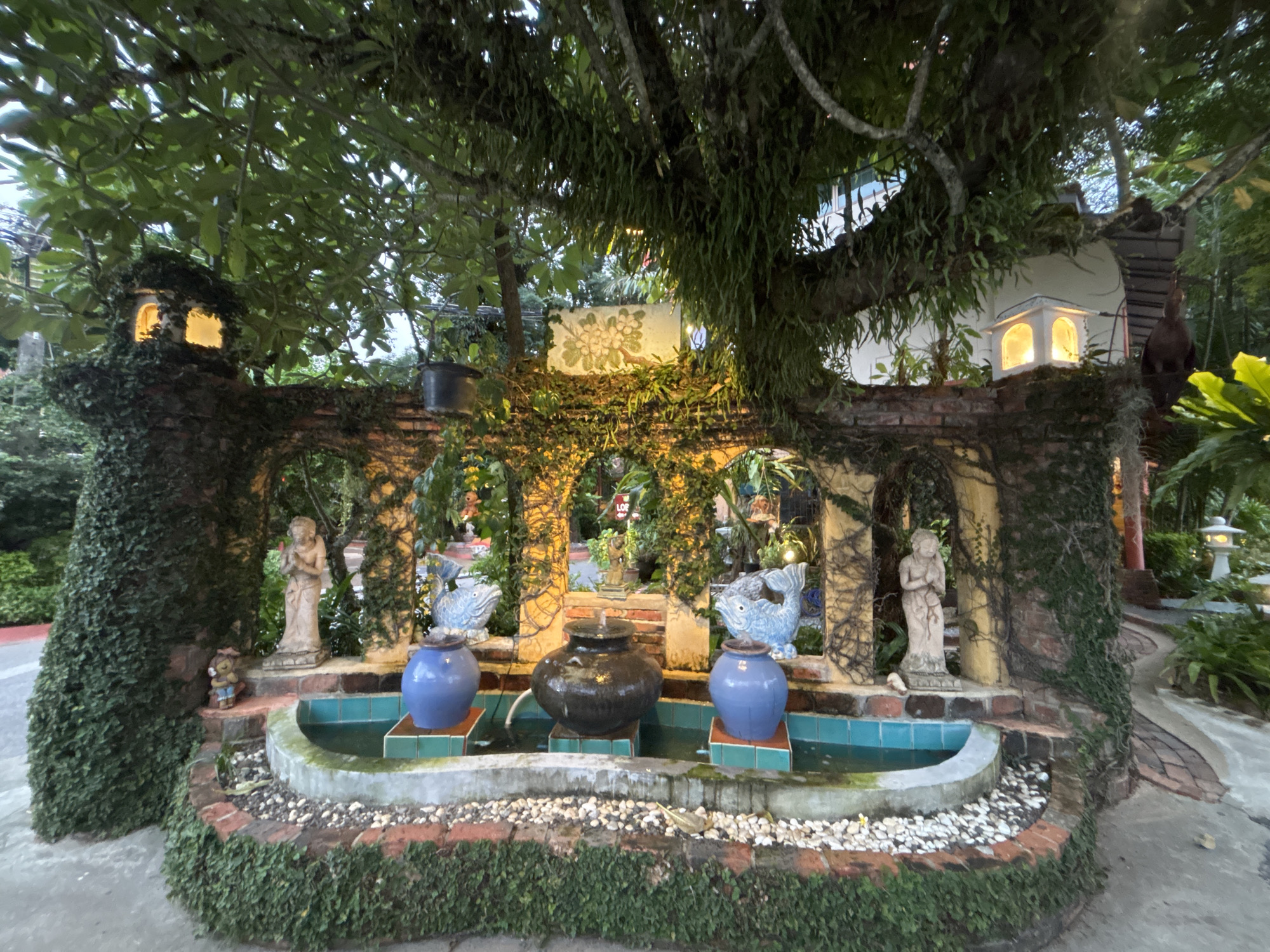 The image captures an outdoor fountain area in Chalong, Thailand, likely in a garden or courtyard setting. The fountain is a tiered structure constructed with brick and stone, adorned with lush green ivy and moss. It features a central, dark-colored pot as the main water feature, flanked by two vibrant blue ceramic vases. Above these, two decorative blue ceramic fish are positioned, suggesting a thematic element to the fountain.
Statues are interspersed within the fountain's design. On the left, a small gnome-like figurine stands near a larger, beige statue of a figure with clasped hands, possibly a representation of a deity or spirit. A similar beige statue of a standing figure is visible on the right side of the fountain.
The background reveals a brick wall covered in vines, with arched openings that suggest walkways or alcoves. Lit structures, resembling small lanterns or decorative lights, are visible on top of the wall, casting a warm glow. A large tree with abundant foliage overhangs the scene, its branches and hanging vines contributing to the natural, somewhat overgrown ambiance. The ground surrounding the fountain is a mix of paved pathways and gravel. The overall scene conveys a sense of tranquil, decorative outdoor space.