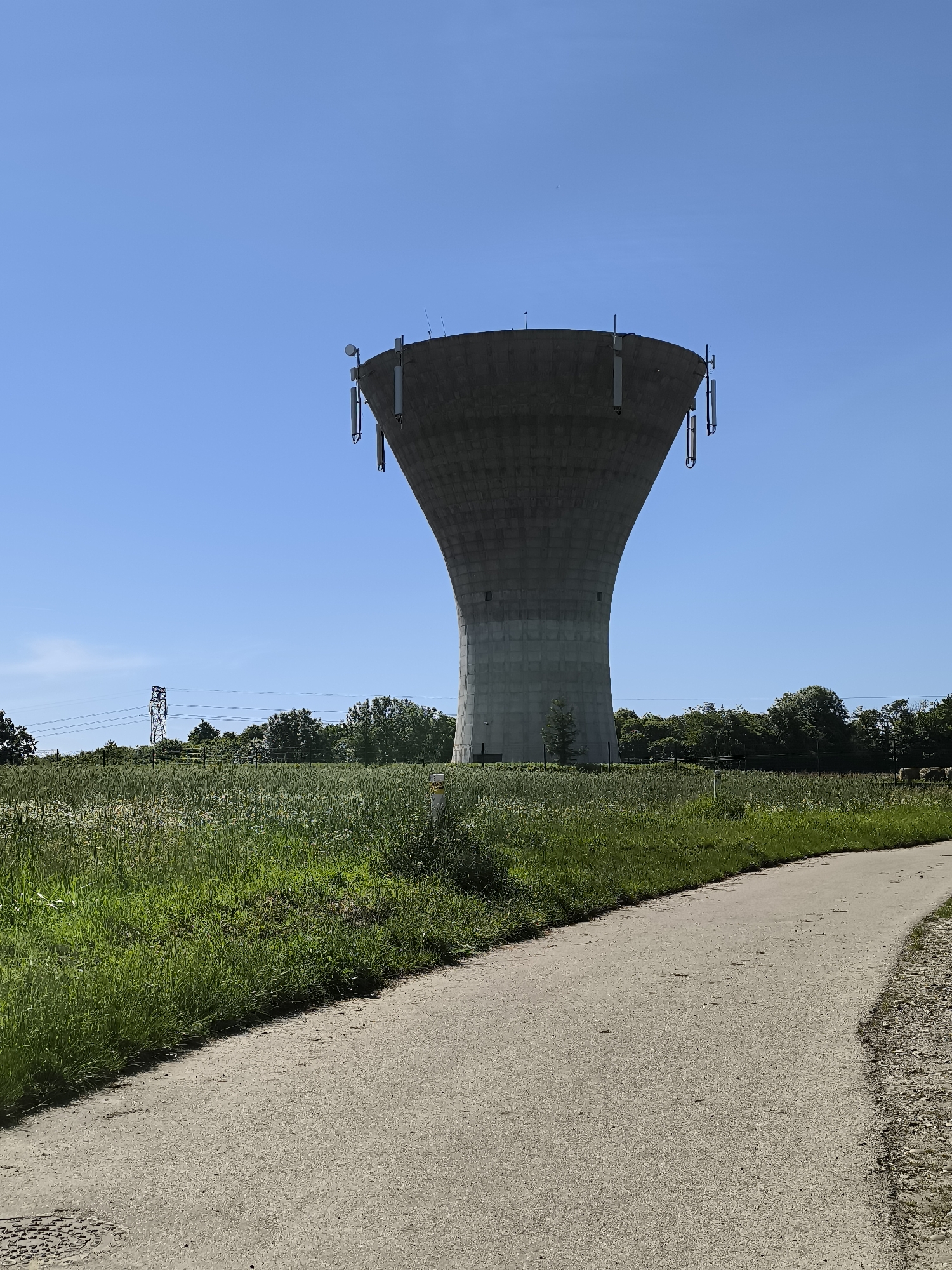 The image captures a water tower in Chaponnay, France, set against a clear blue sky. The tower is light gray with multiple antenna mounted at its top, visible against the backdrop of the sky. The landscape includes a path, tall green grass, and trees, creating a contrast to the industrial architecture of the water tower. An electrical tower is visible in the background.