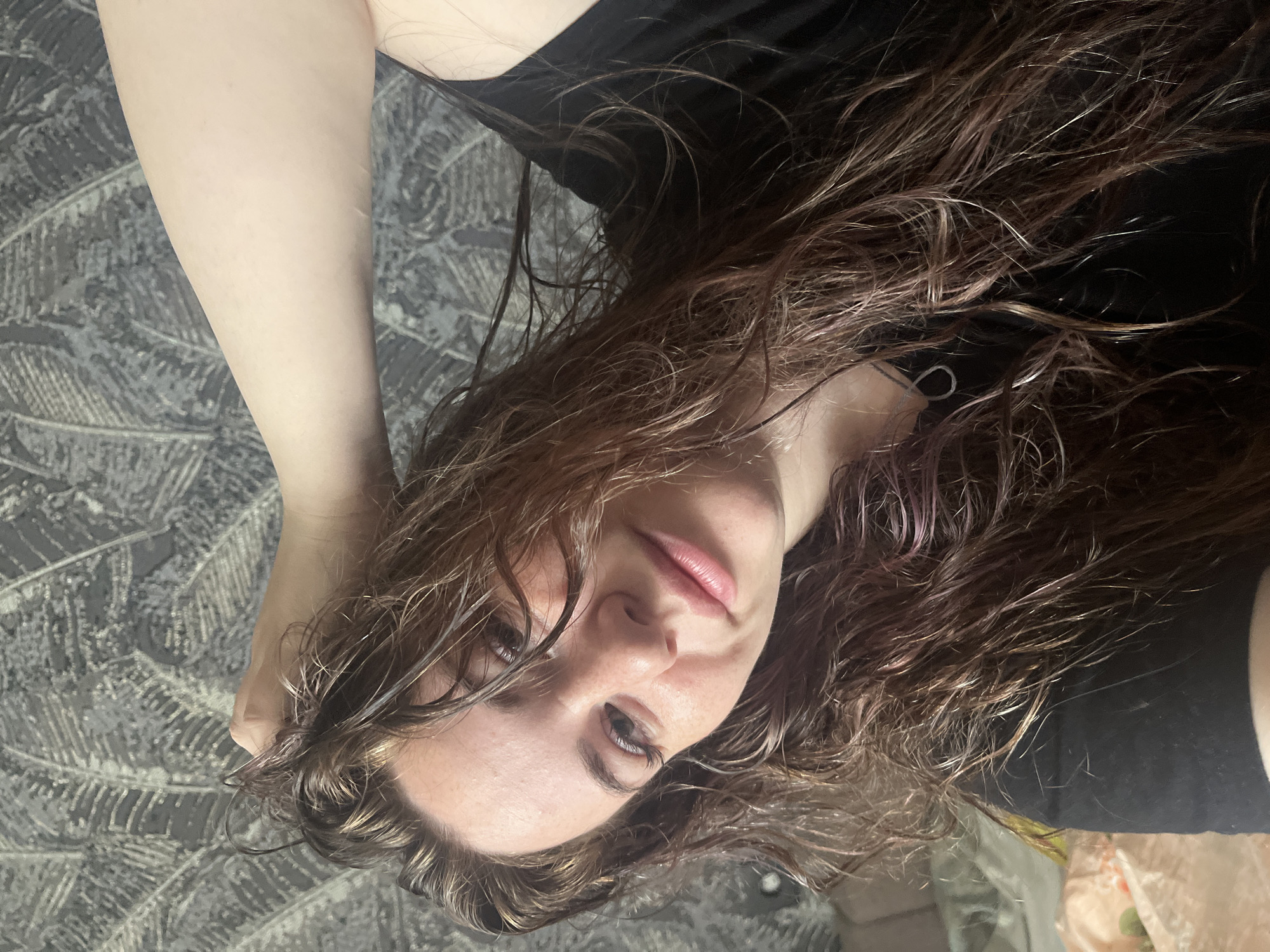 A young woman with wet, wavy brown hair is lying on a patterned gray surface. She is wearing a black shirt. Her face is visible, with light skin, brown eyes, and pink lips. The location cannot be determined to be Chaikovsky, Russia from the image.
