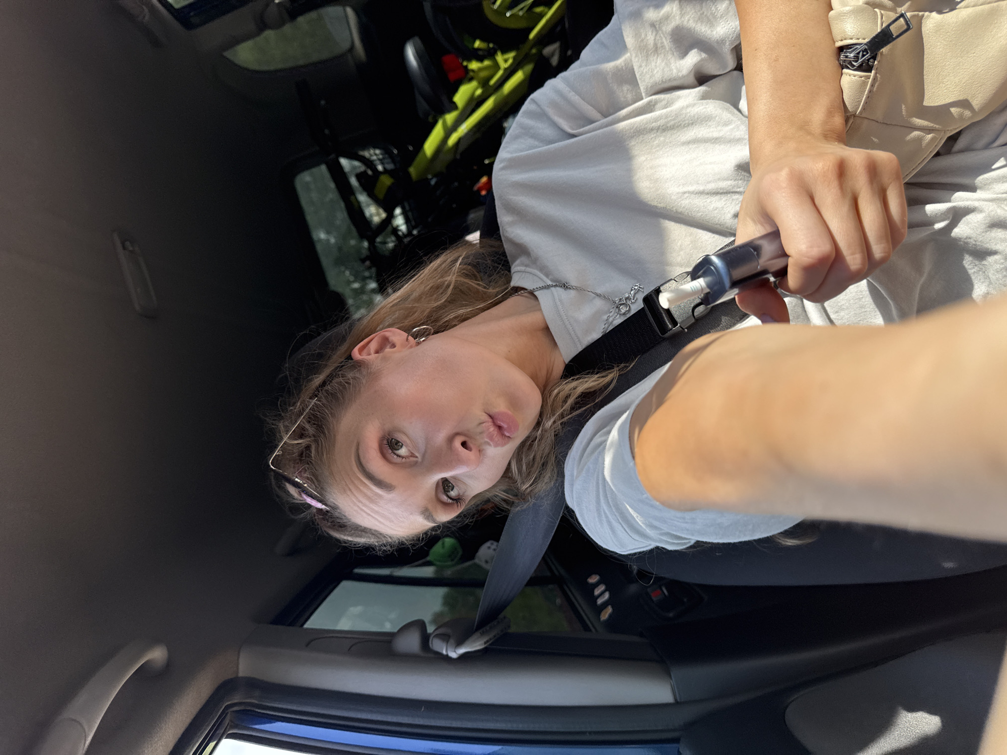 A young woman is in the passenger seat of a vehicle. She is looking at the camera with her lips pursed and is holding a vaping device. She is wearing a light-colored t-shirt and a silver necklace. A beige purse is visible on her lap. In the background, there are bicycles in the back of the vehicle. The vehicle appears to be in a rural area with trees visible through the window. The image was taken on a sunny day. The city of Perm, Russia, is not visible in this image.